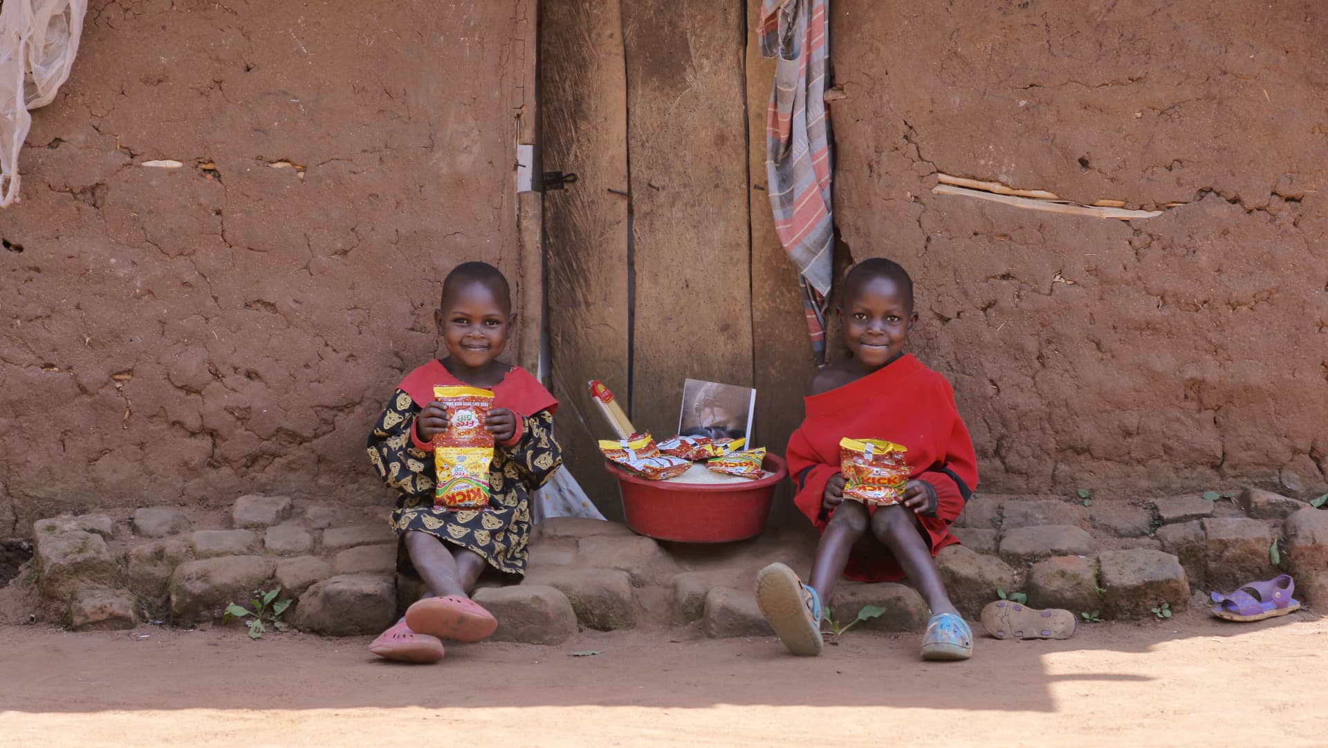 Two children in red outfits sitting against a mud-brick wall with a food bowl and a photo card