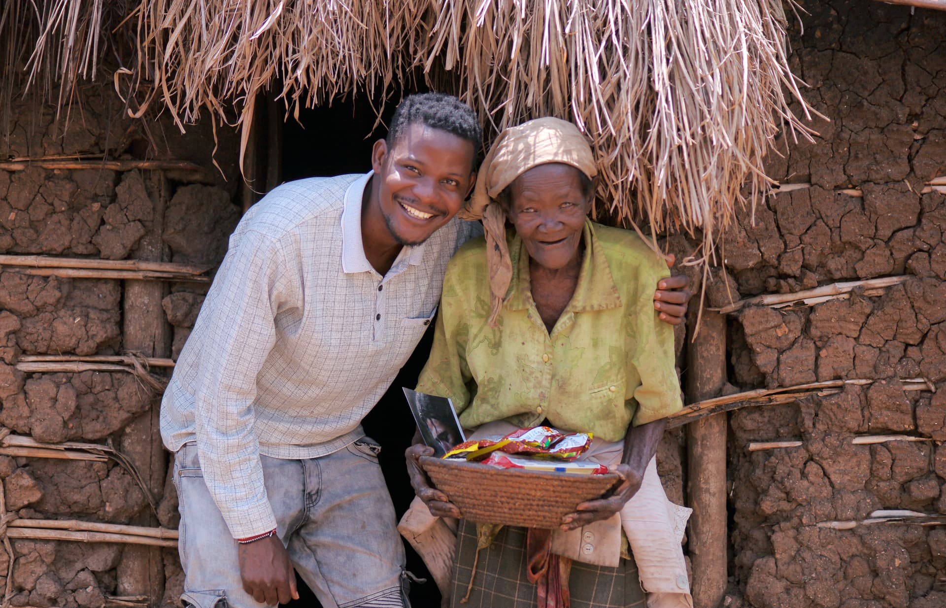 Elderly man and woman stand together smiling outside a mud-brick hut, holding a food gift basket.