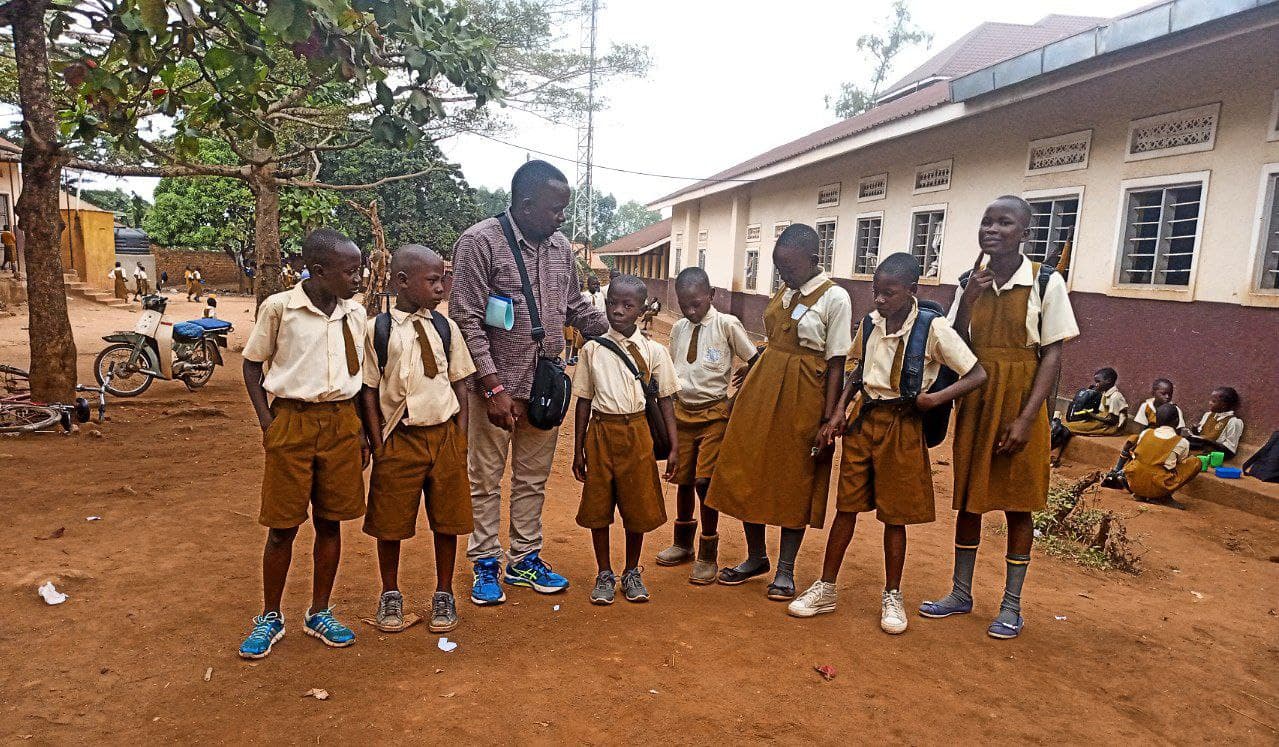 Mashariki Children's Home schoolchildren in brown uniforms gather outside their school building with a staff member.