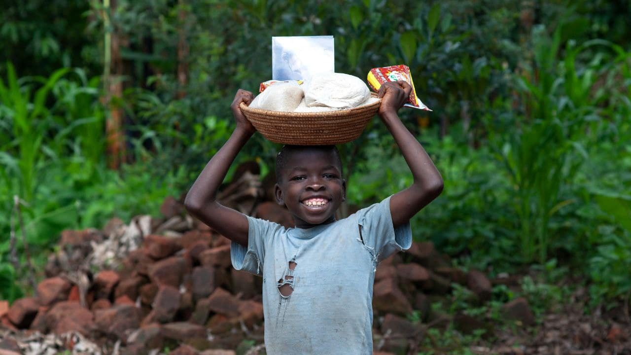 Boy carrying a white basin of food supplies on his head while walking through greenery