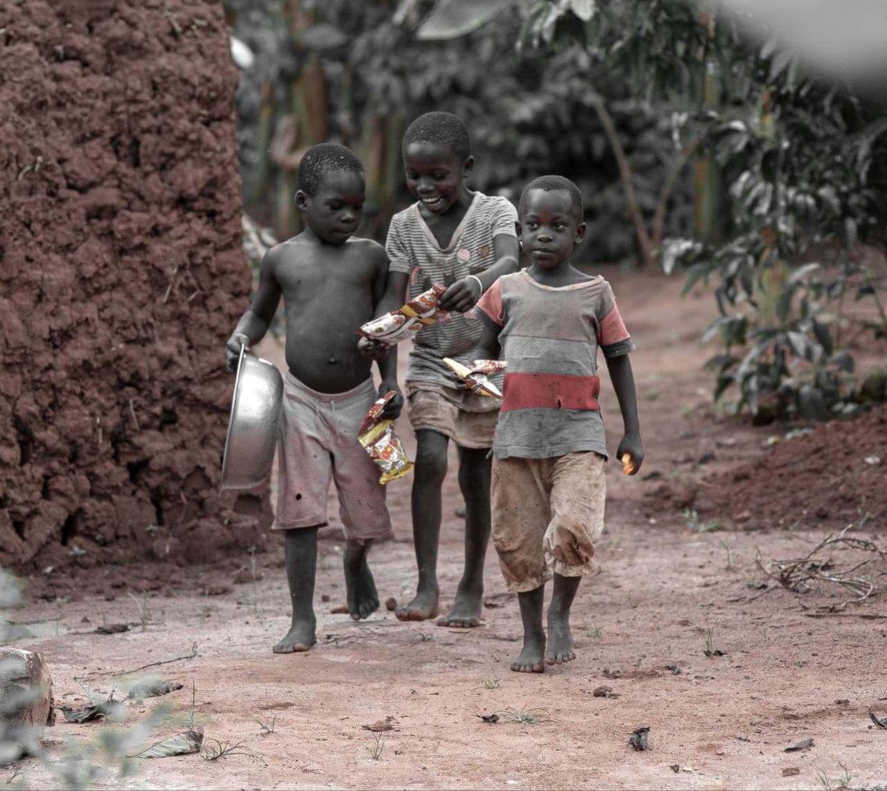 Three children walking together along a dirt path near a mud-brick building, one carrying a bag