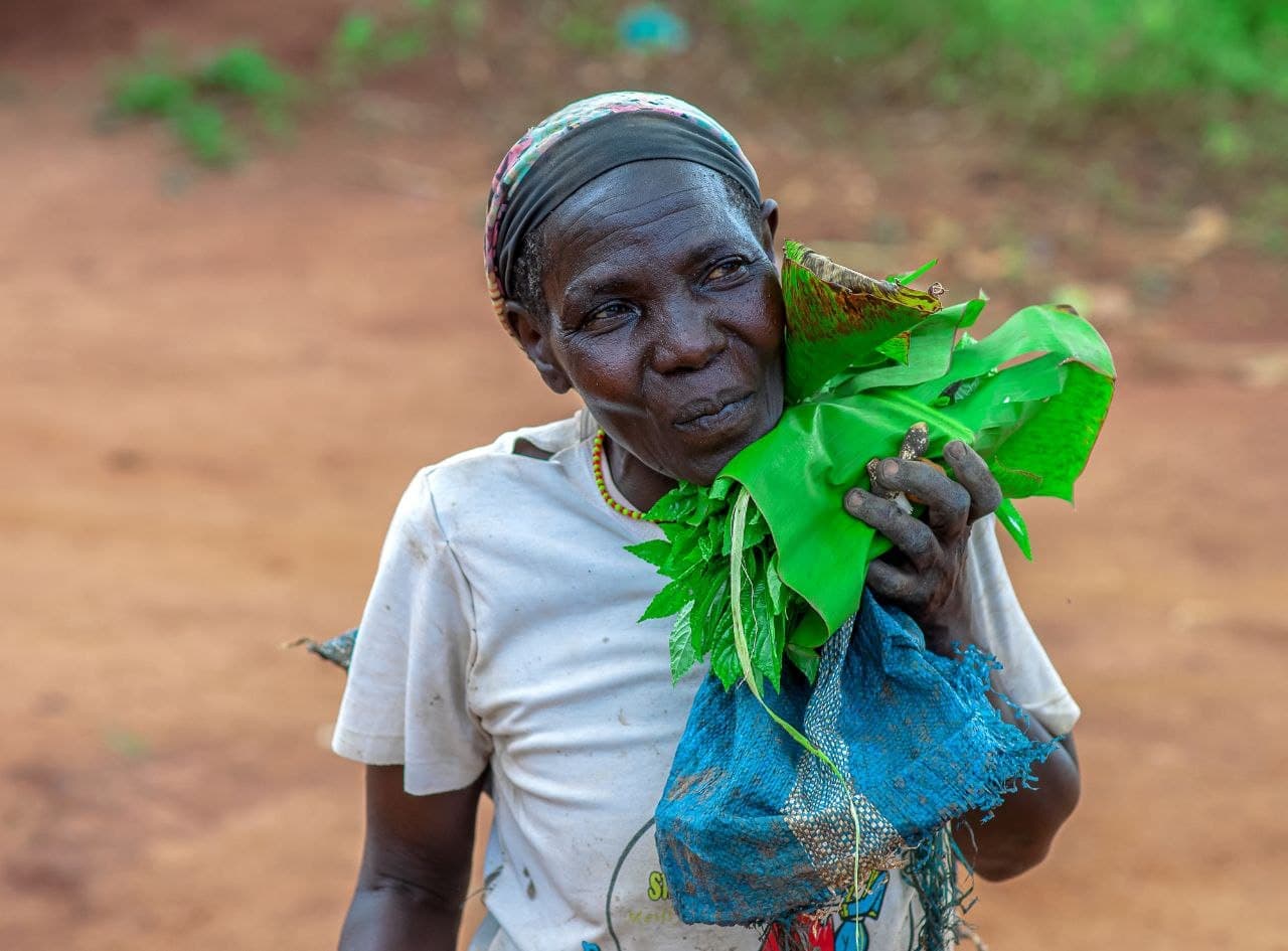 Elderly woman in white shirt and headscarf holding green leafy vegetables and a blue mesh food bag