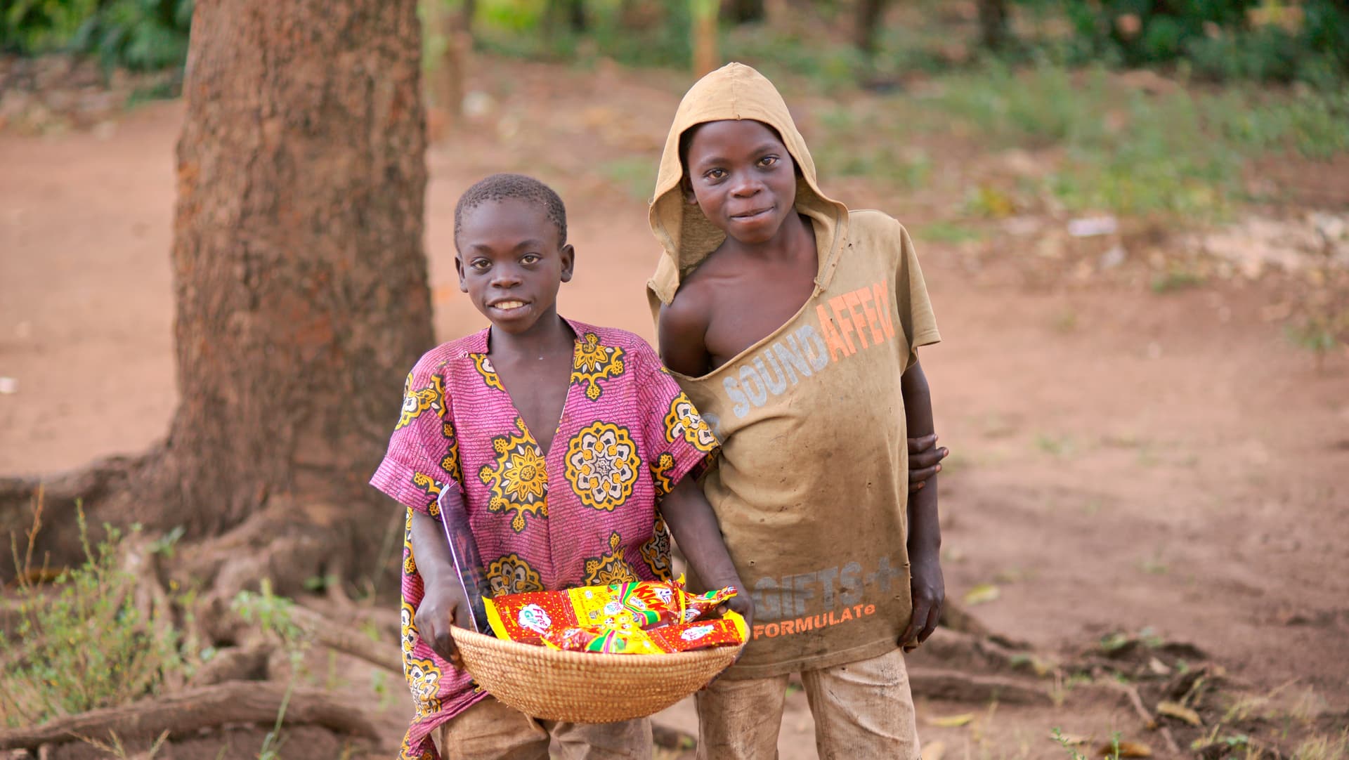 Woman and child standing together outdoors holding a basket of food items under a tree