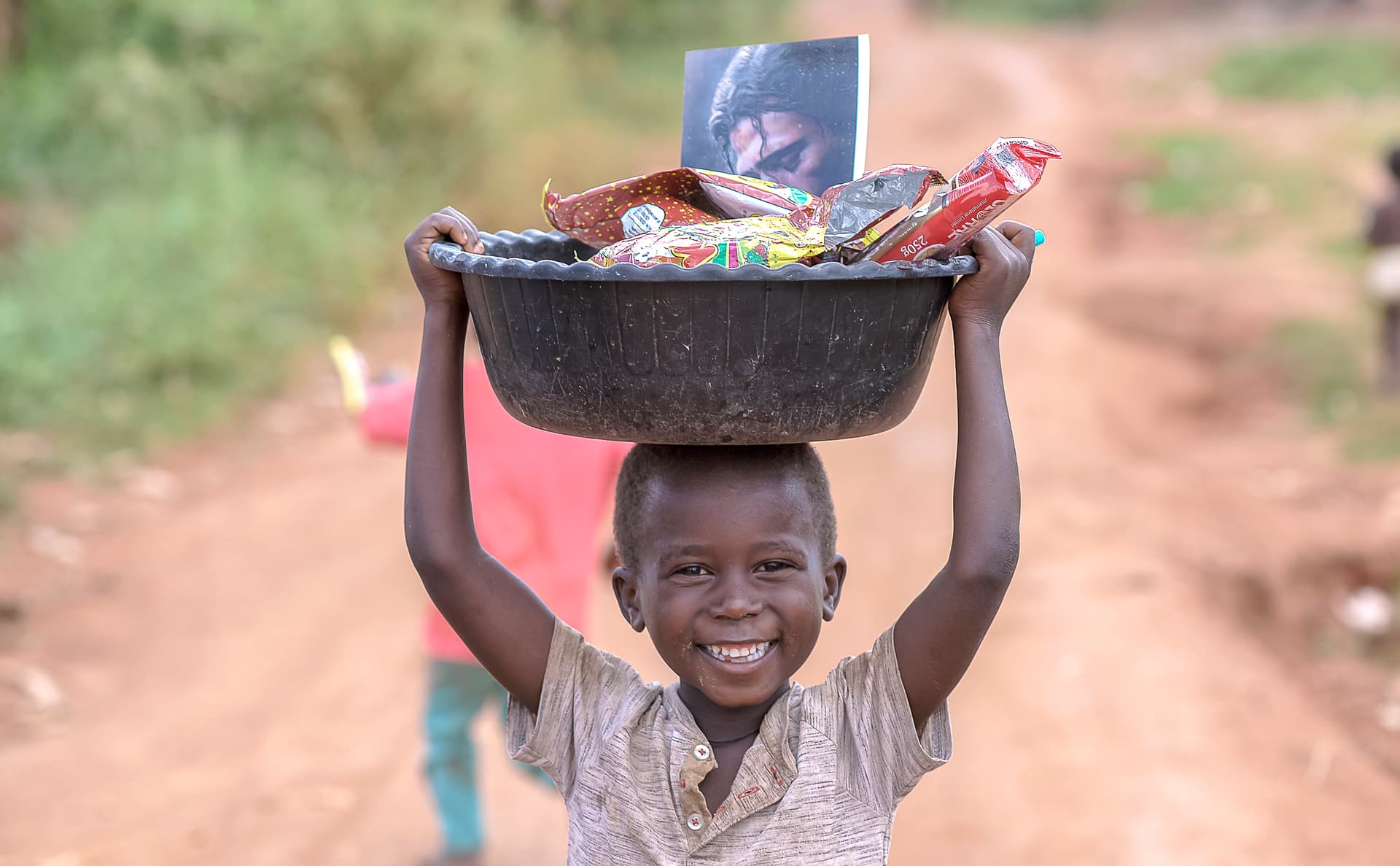 Boy carrying a black basin of food packets on his head while walking down a dirt road