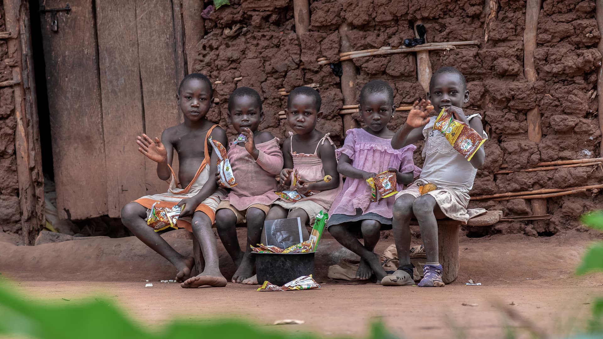 Row of young children in matching pink outfits sitting together against a mud-brick wall