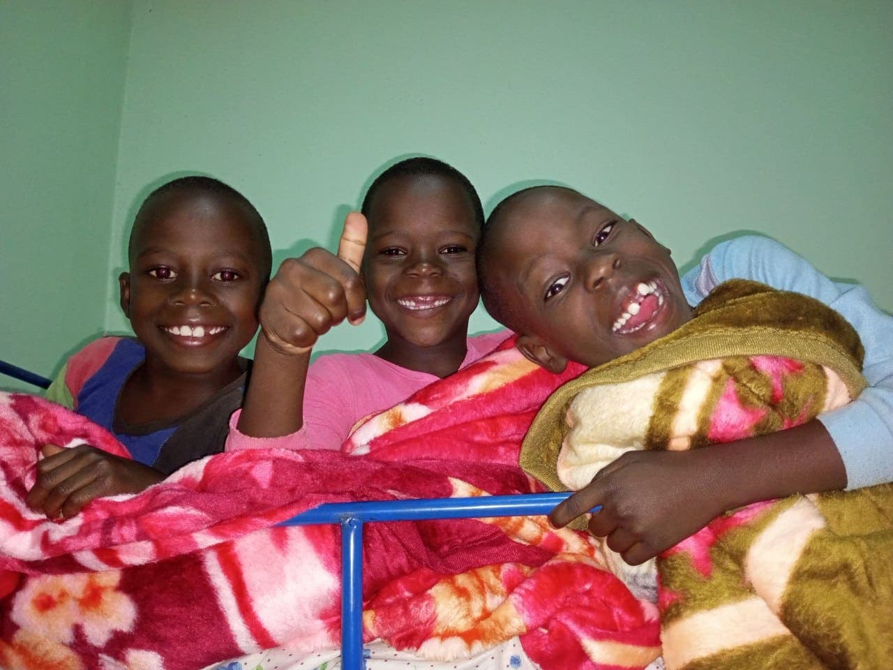 Three children lying in bed together giving thumbs up under a colorful floral blanket at Mashariki Children's Home