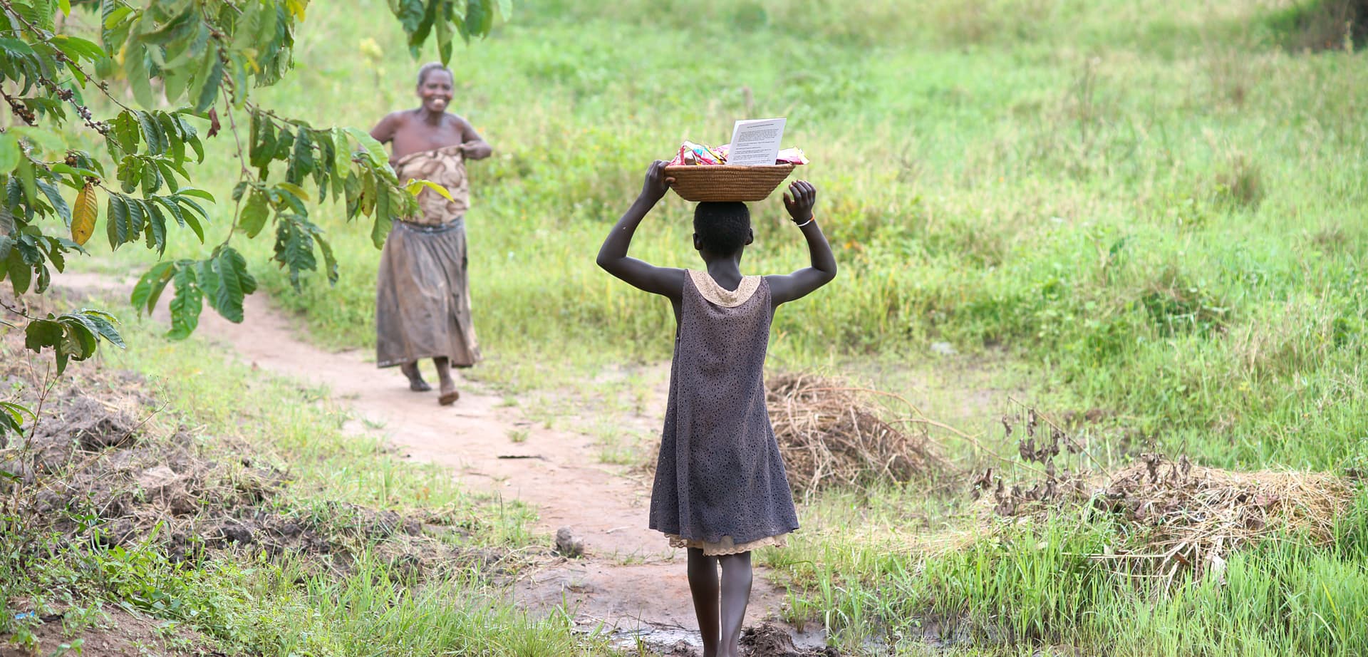 Two women or girls in headwraps carry items on their heads while walking along a dirt path.