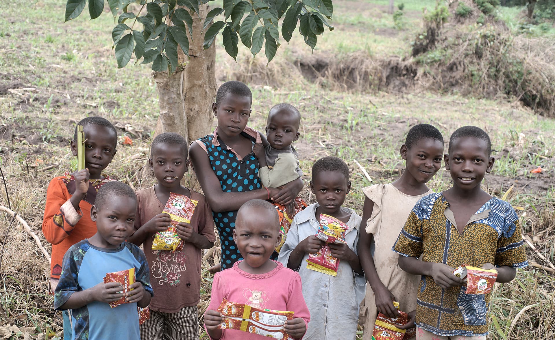 A group of children and a woman stand together under a tree outdoors, some holding gospel tracts.