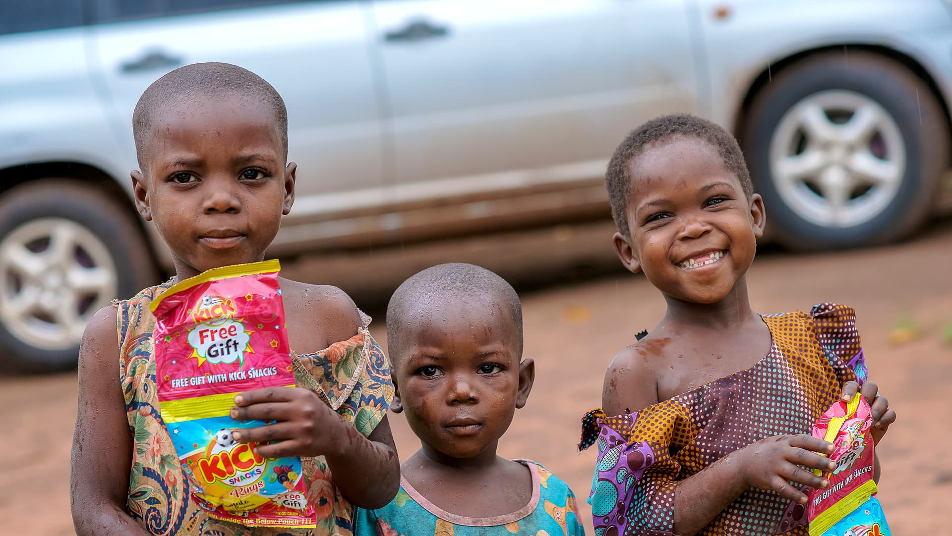 Two smiling children hold colorful food packages with a car parked behind them.