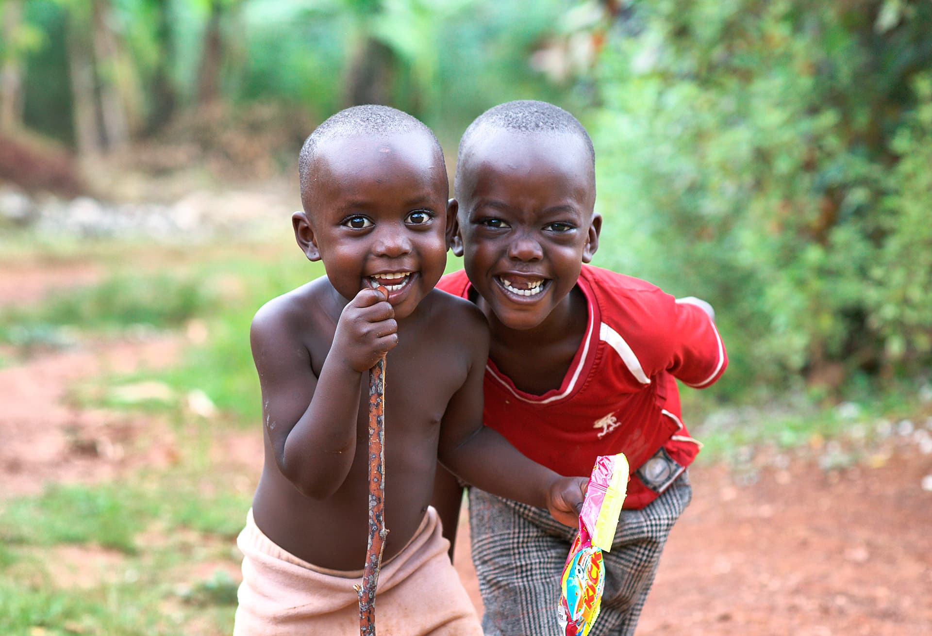 Two boys stand close together outdoors, smiling and gesturing toward the camera.