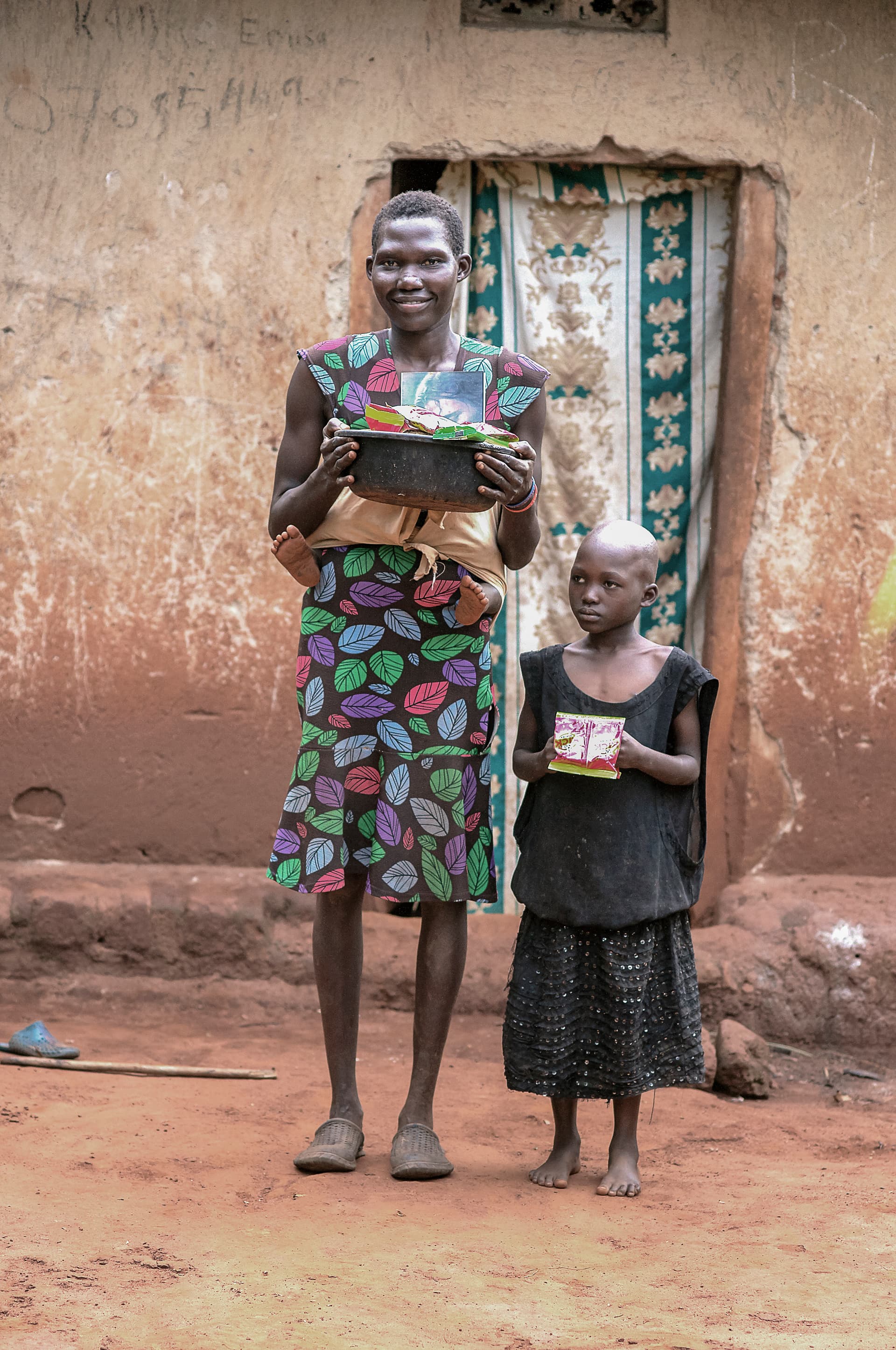 Woman stands in a mud-brick doorway holding food items beside a young girl holding a small card.
