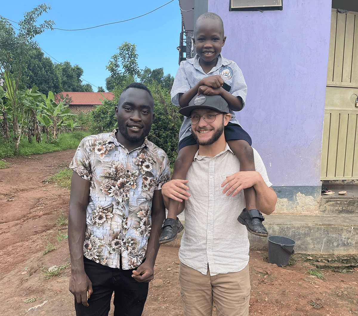 Man carries a young boy in a school uniform on his shoulders beside another man outside a purple building.