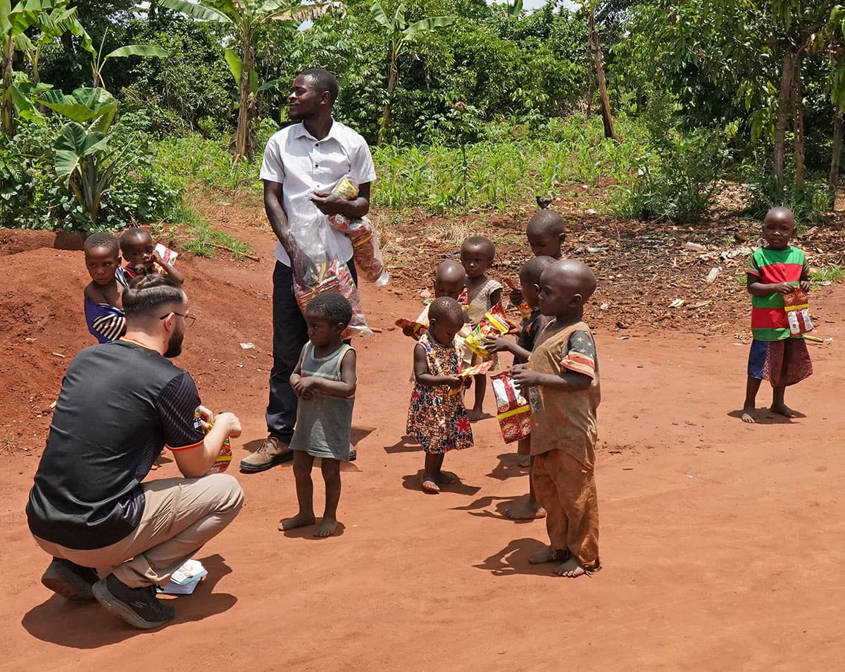 A JFM team member kneels to hand food to children gathered on a red dirt path.