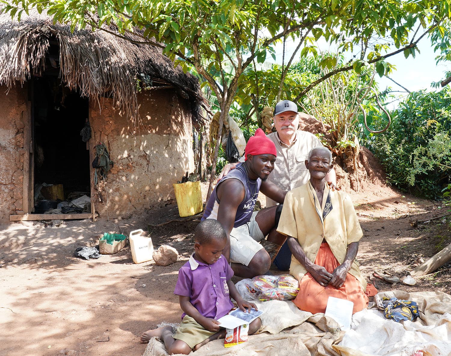 Bill and JFM team members kneel with two children outside a mud hut, food items on the ground.