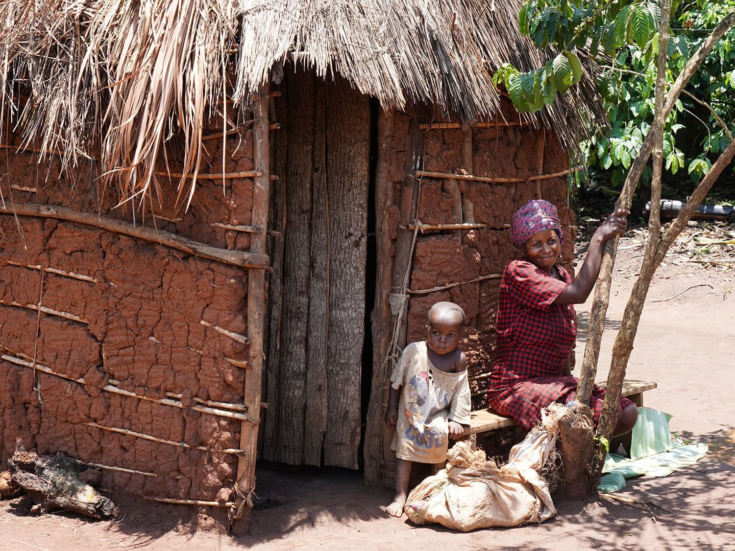 Elderly woman in a headscarf sits outside a mud-and-thatch hut beside bundled sacks.