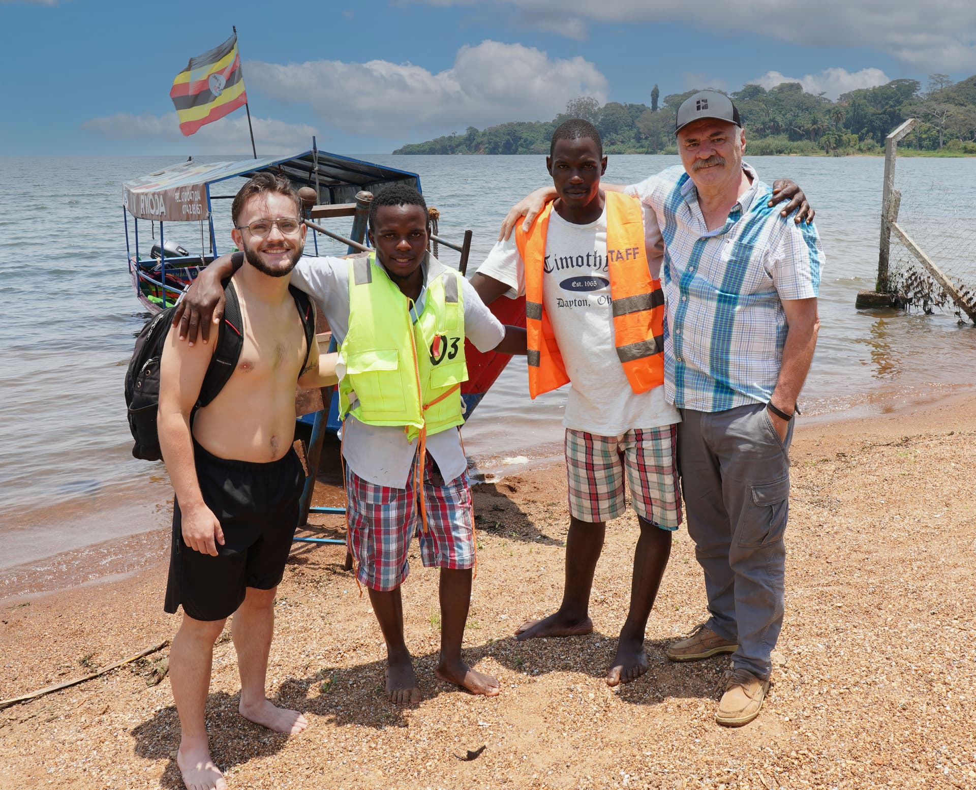Four men stand together on the shore of Lake Victoria, two wearing JFM reflective vests.
