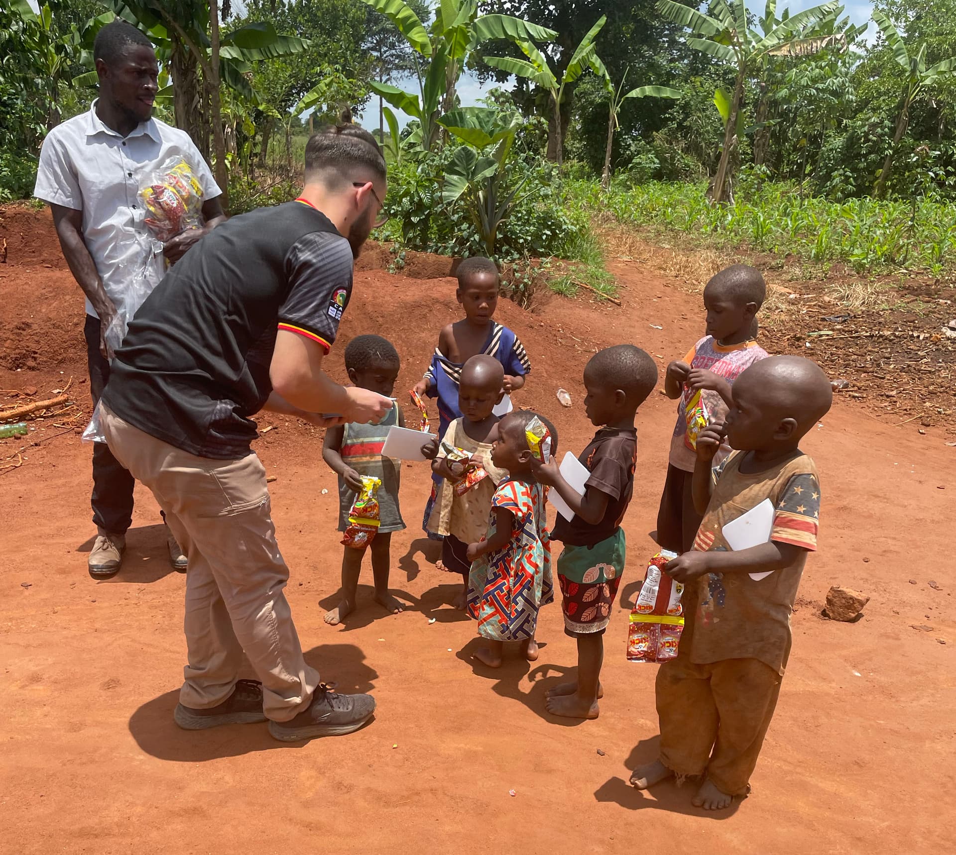Man crouches on a dusty road handing food or tracts to a line of young children.