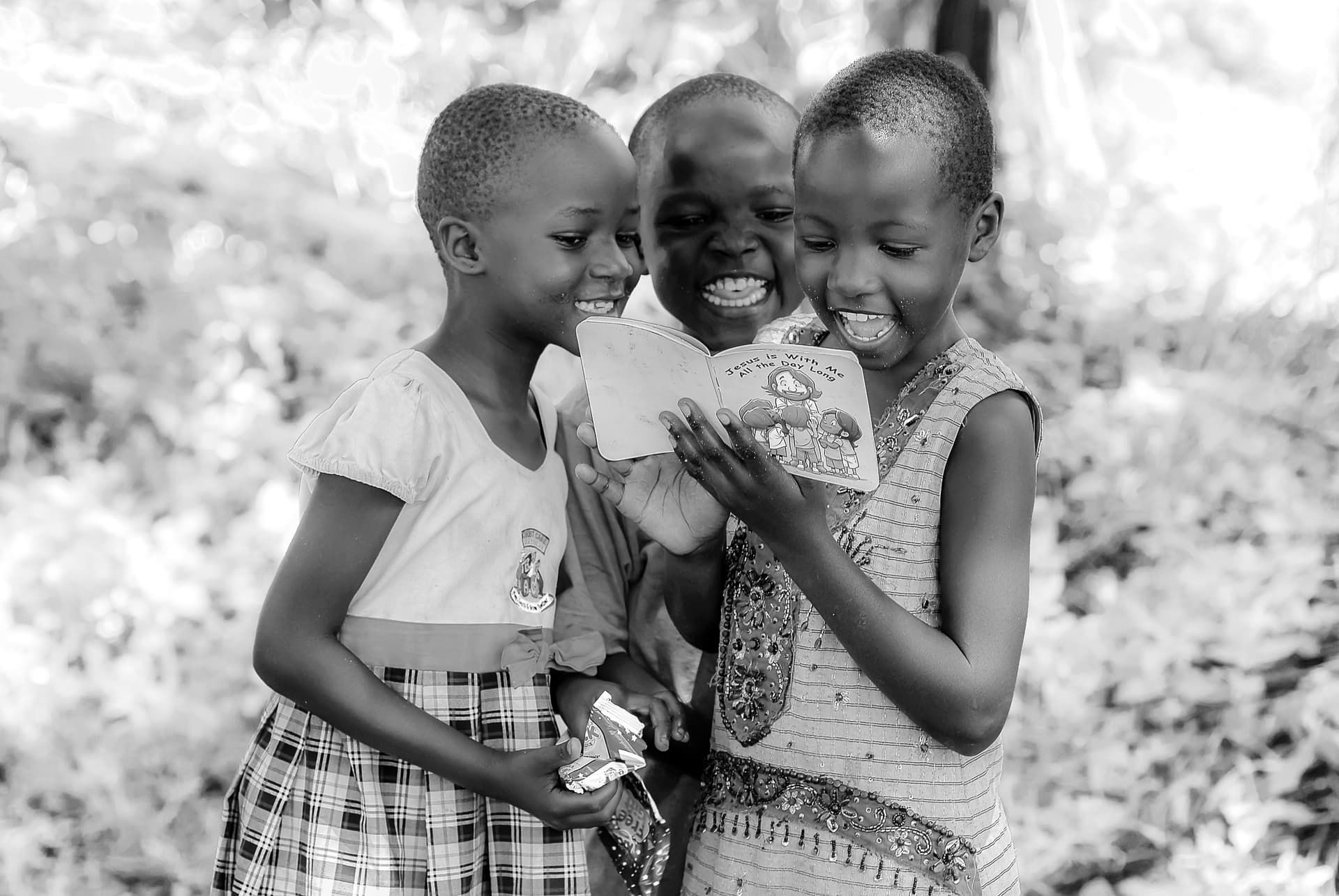Black and white photo of three young children laughing together, one holding a small children's Bible booklet.