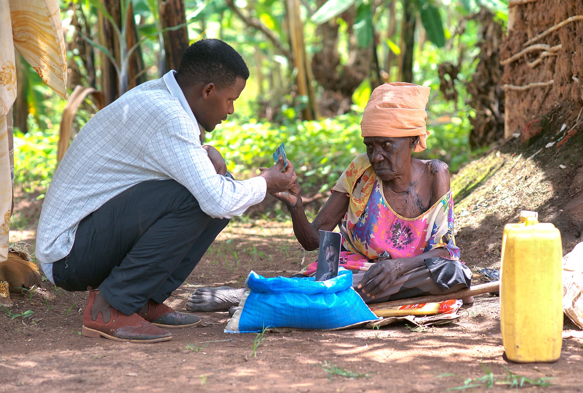 A JFM team member kneels to hand Elder Atim Justine a food package as she sits outside her hut.