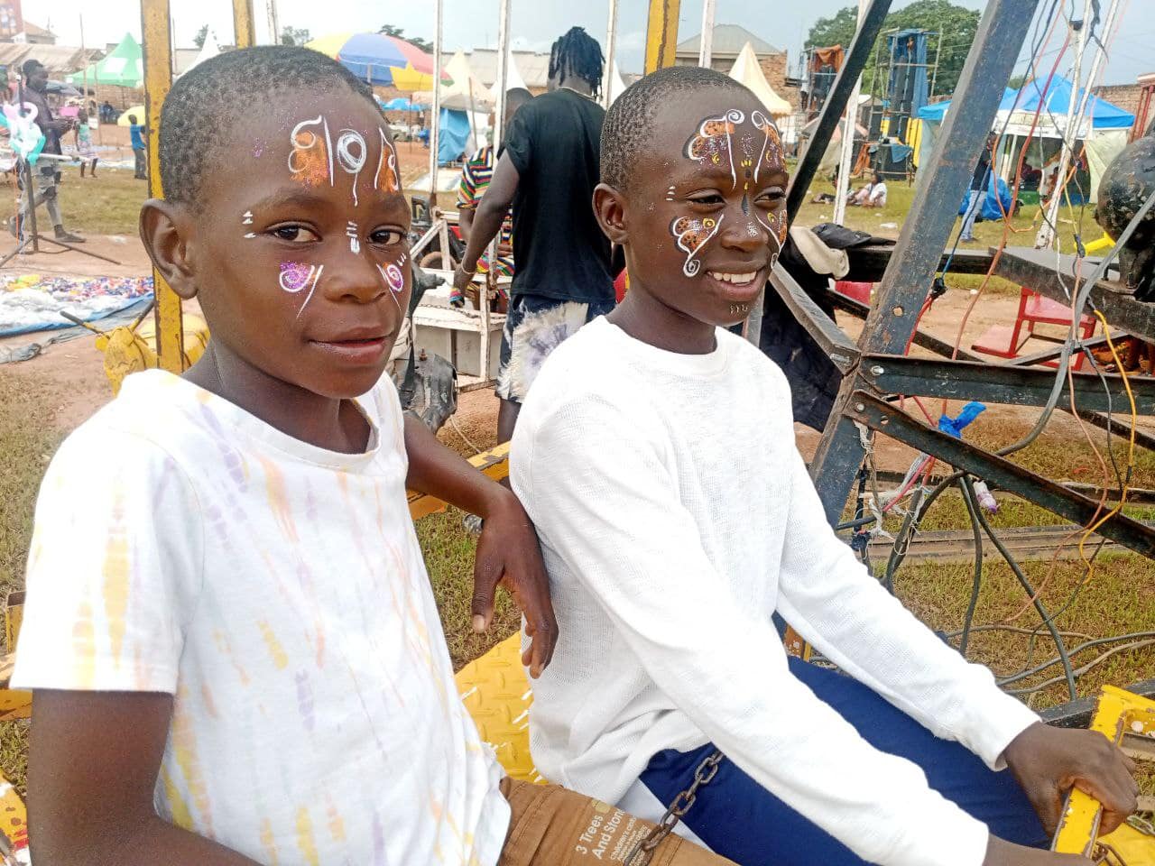 Two Mashariki boys with painted faces sit on a ride at an amusement park.