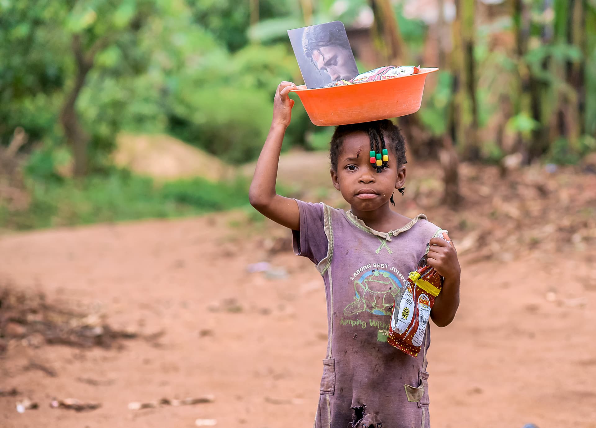 Cherry balances a food container on her head while standing on a dirt path.