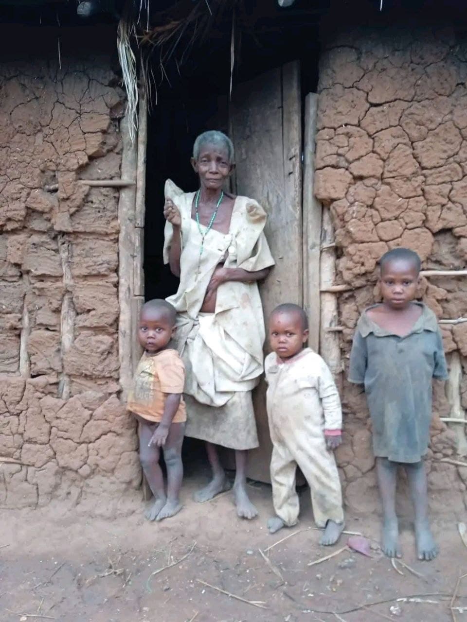 Magana, an elderly woman, stands in the doorway of her mud and thatch hut with three children.