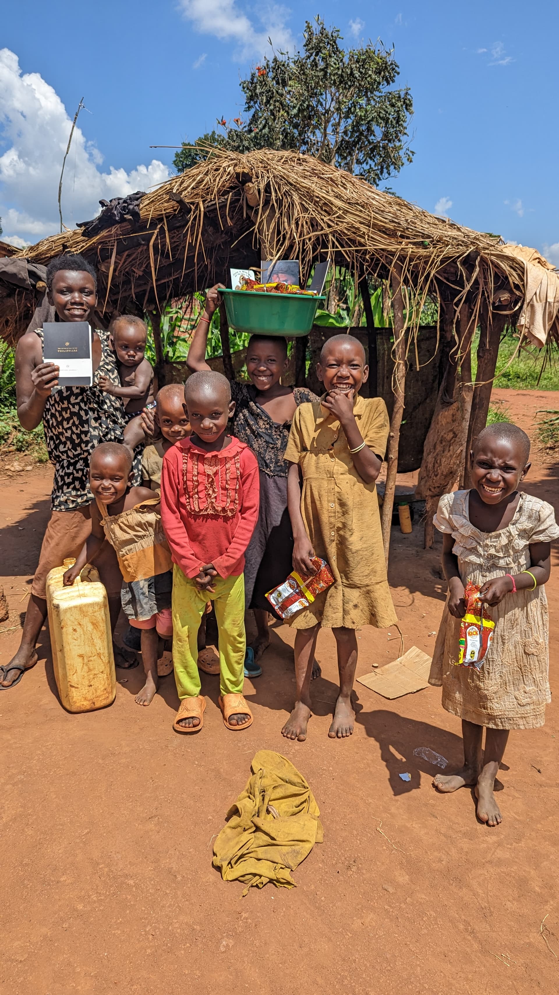 A group of children stand outside a thatched hut holding food packages.