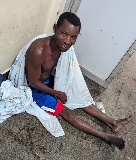 Emma sits on the ground against a wall, shirtless and wrapped in a white cloth, at Miracle Medical Center.