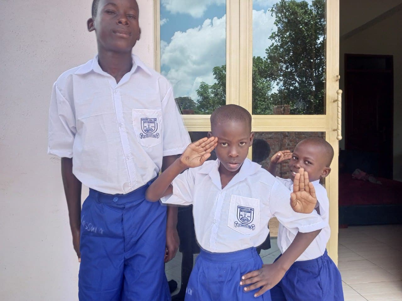 Three schoolchildren in blue and white uniforms stand together indoors, one saluting.