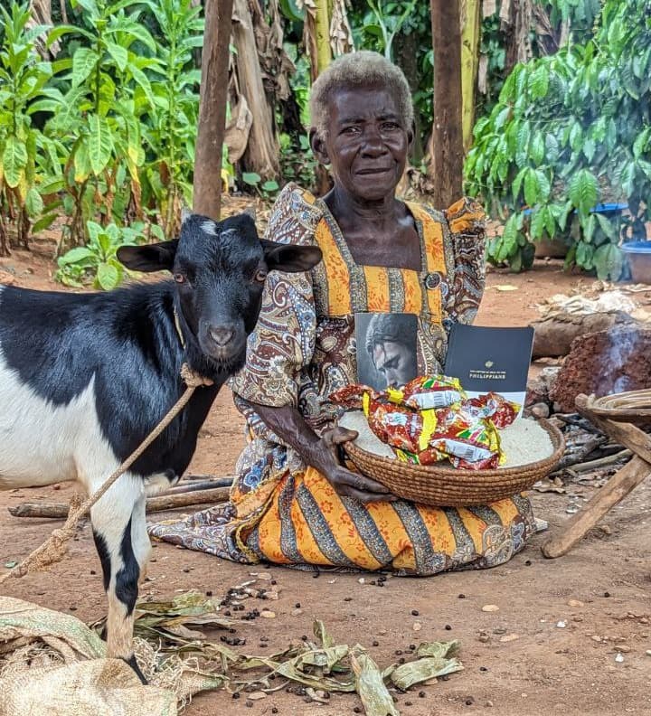 Maria, an elderly woman, sits with her black and white goat and a basket of food gifts.