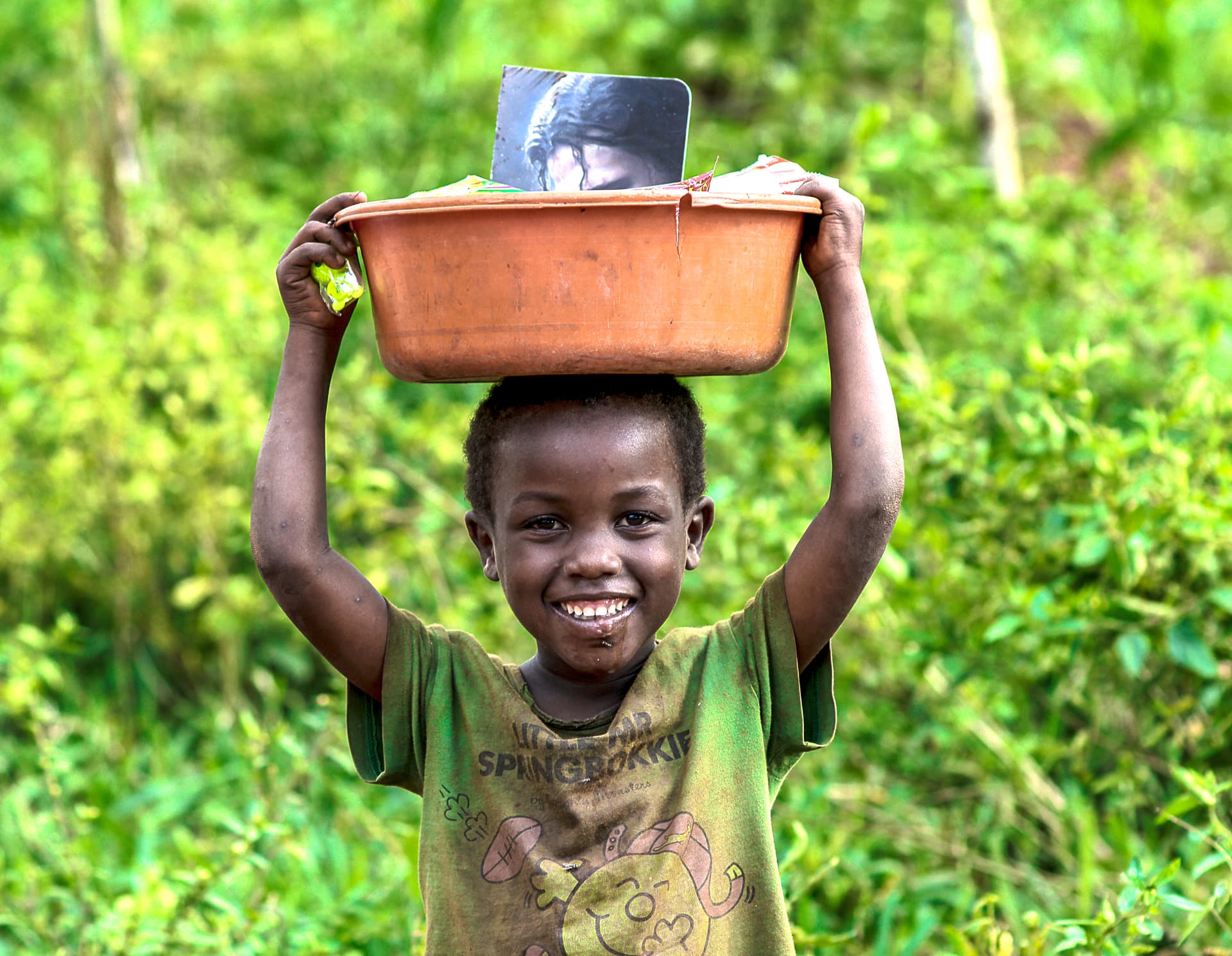 A smiling boy balances a basin loaded with a JFM food gift package on his head.
