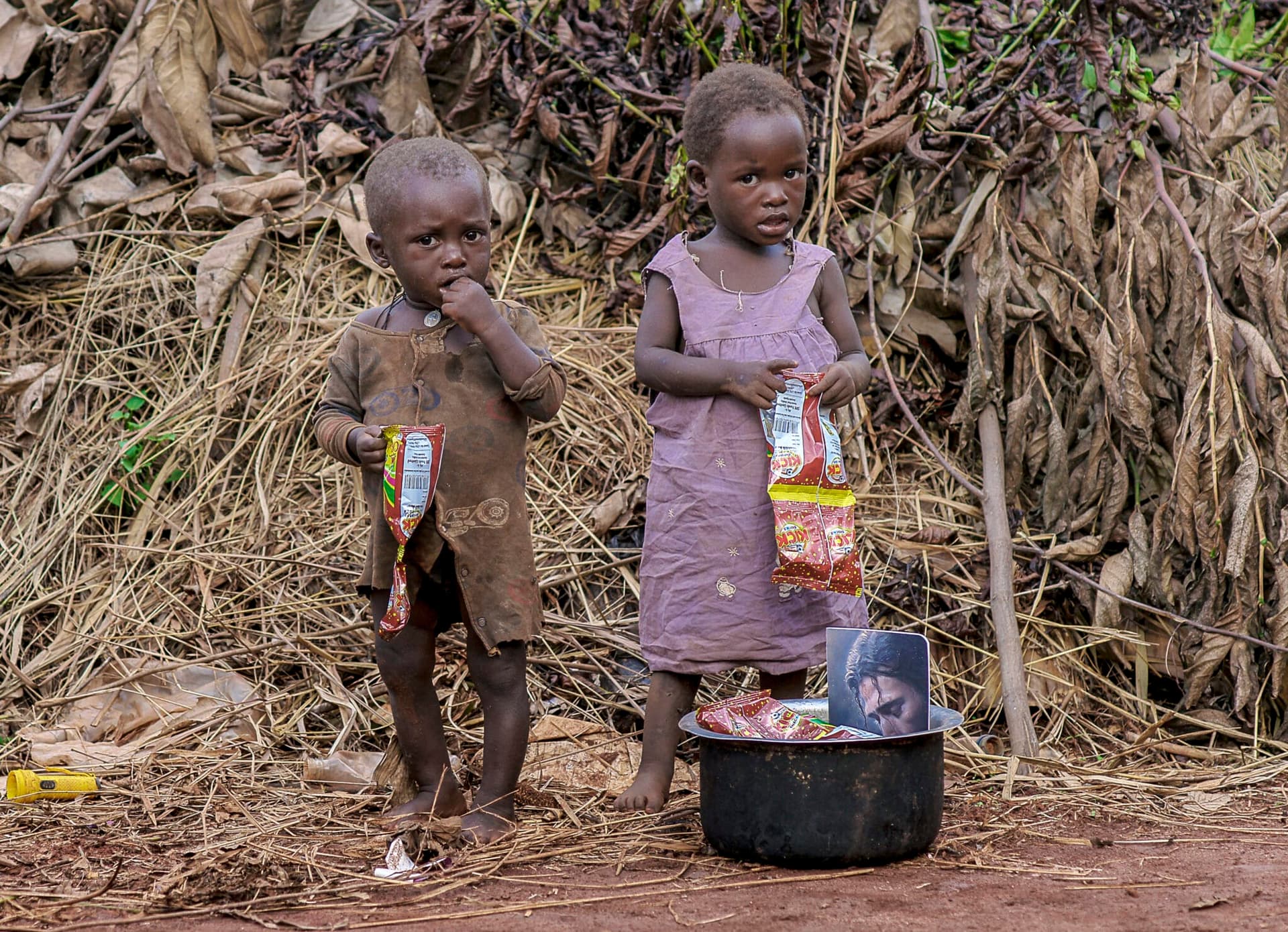Two children stand outdoors near a straw structure, one holding a basin of food items.