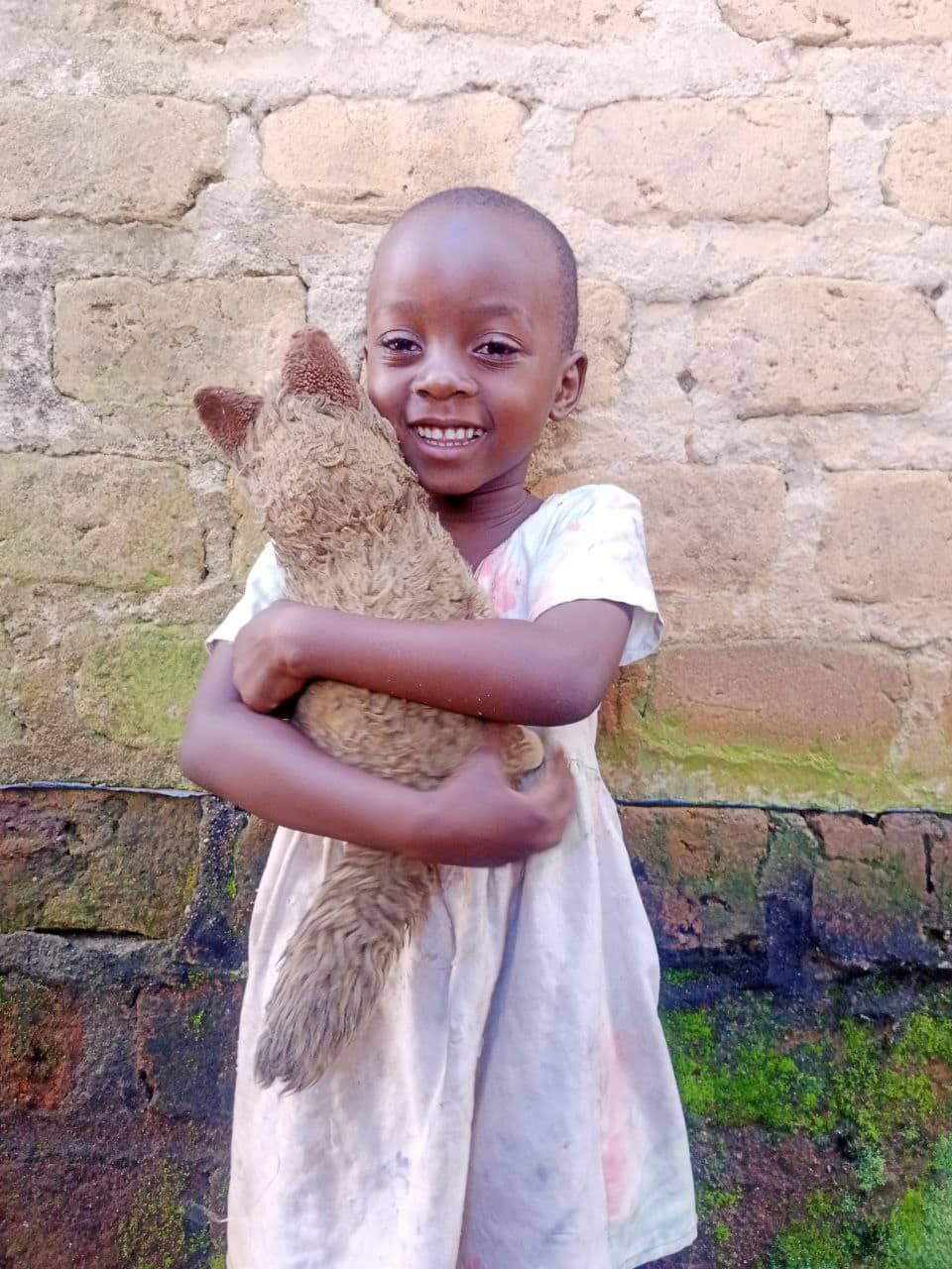 Sharon, a young girl in a white dress, smiles while hugging a stuffed teddy bear against a brick wall.