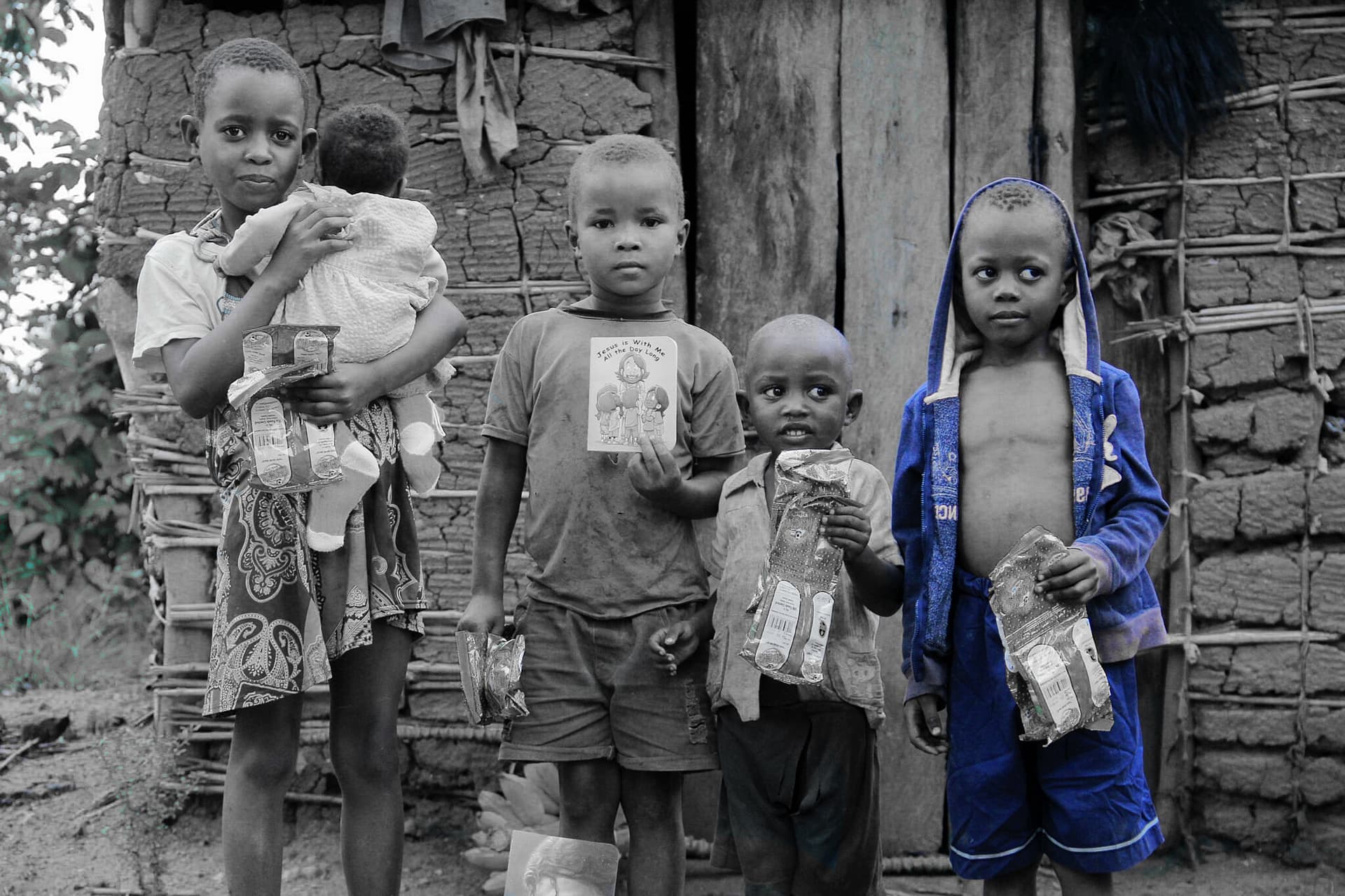 Black-and-white photo of a woman carrying a baby with children standing beside a mud house.