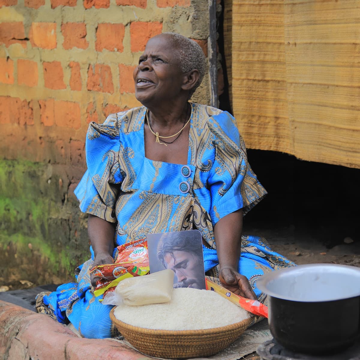 Granny Elizabeth sits outside her home preparing food, with a bowl of cassava flour and a kettle nearby.
