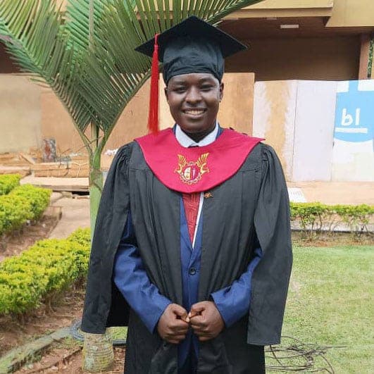 A man in a graduation cap, gown, and red sash stands outdoors after his ceremony.