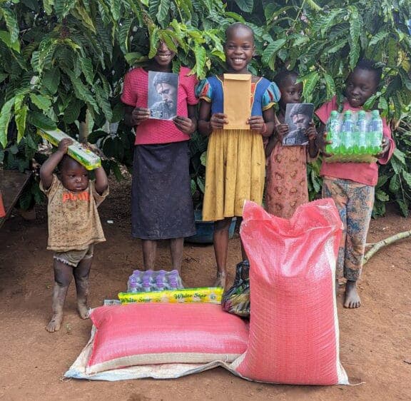 Children stand together holding gospel tracts beside red sacks of Christmas food supplies.