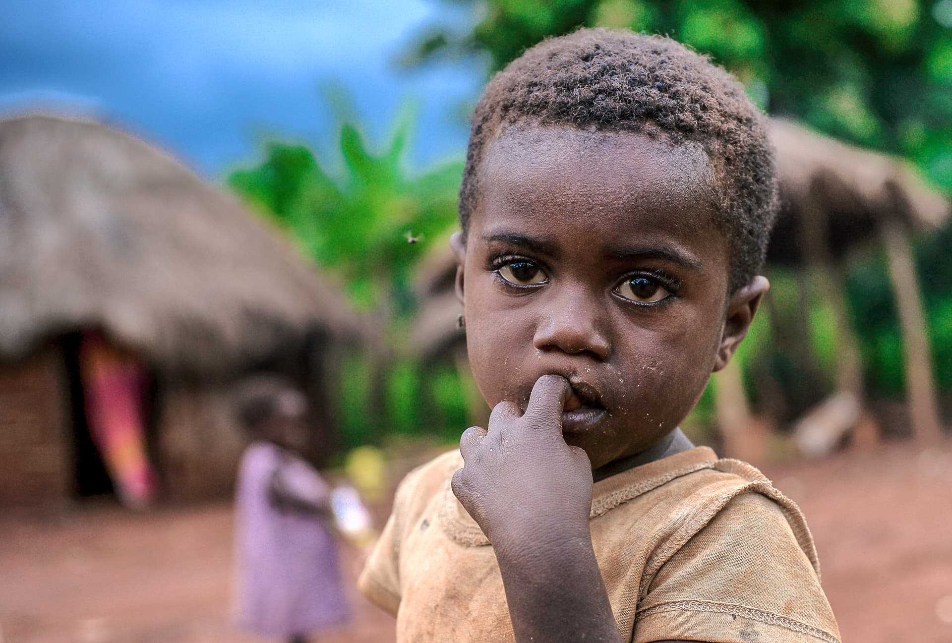 Young boy with a serious expression, finger near his mouth, thatched huts in the background.