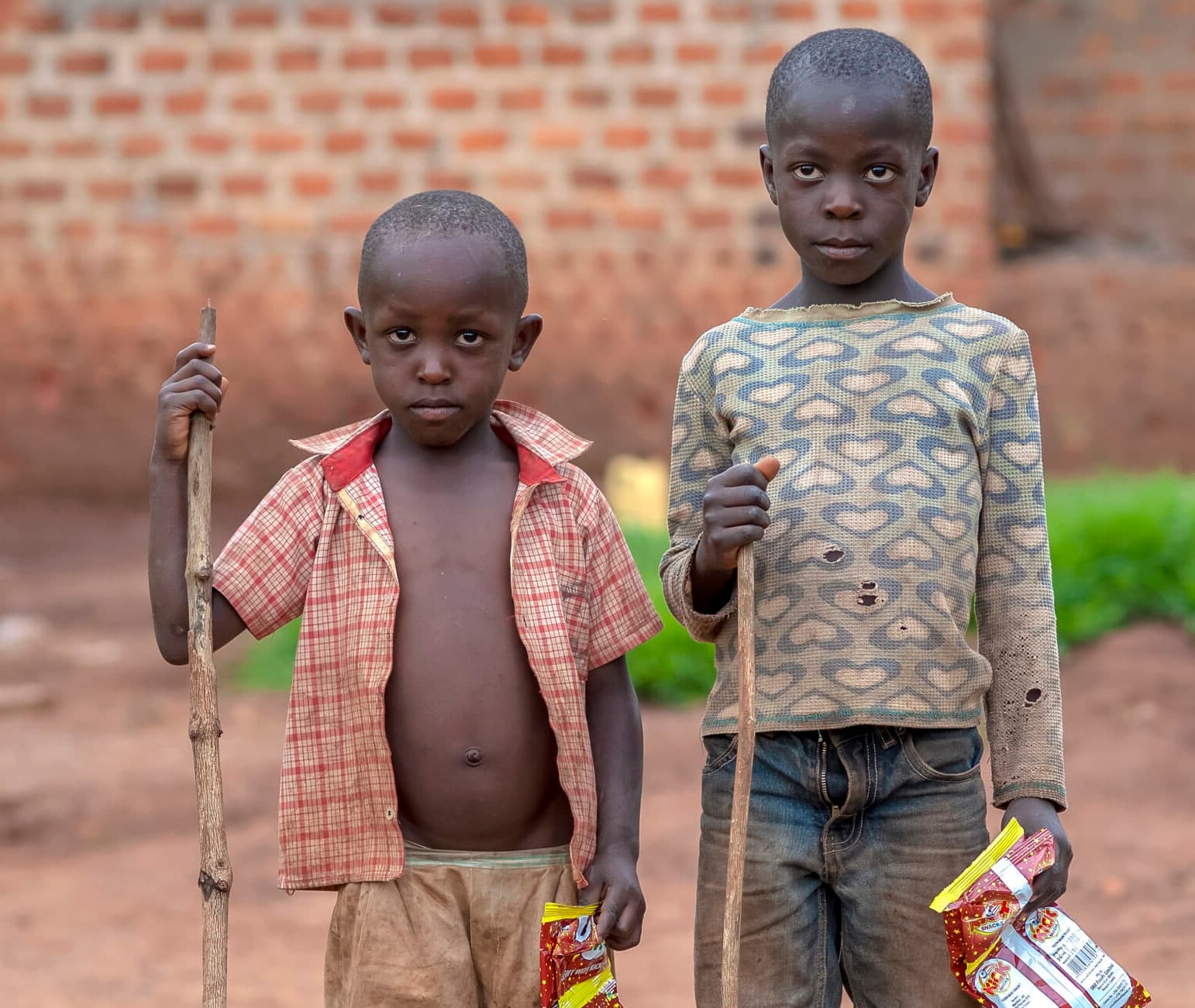Two boys in tattered clothes stand on a dirt path, each holding a walking stick and a small food package.