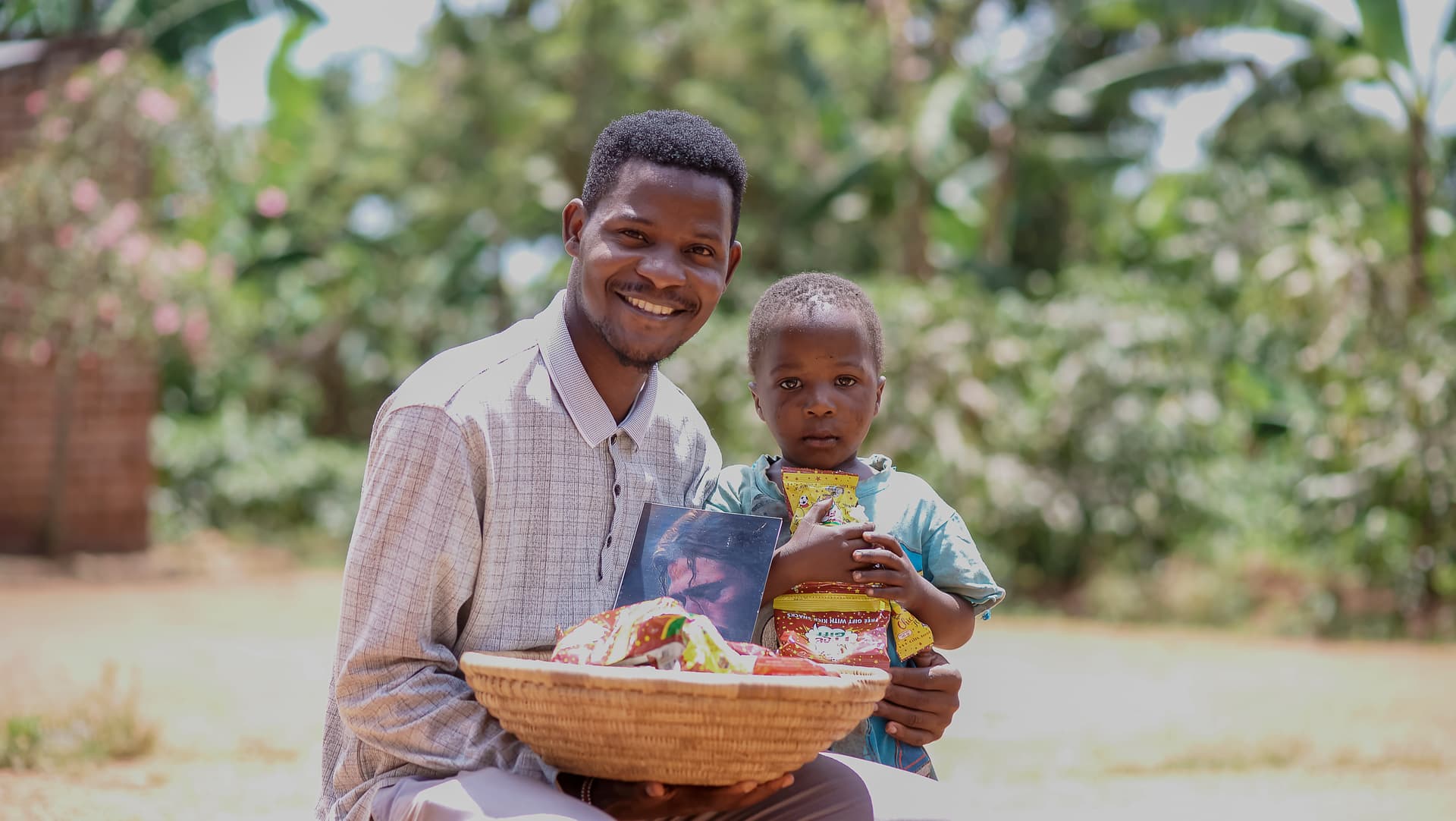 Man sits with a young child beside him, holding a woven basket outdoors.