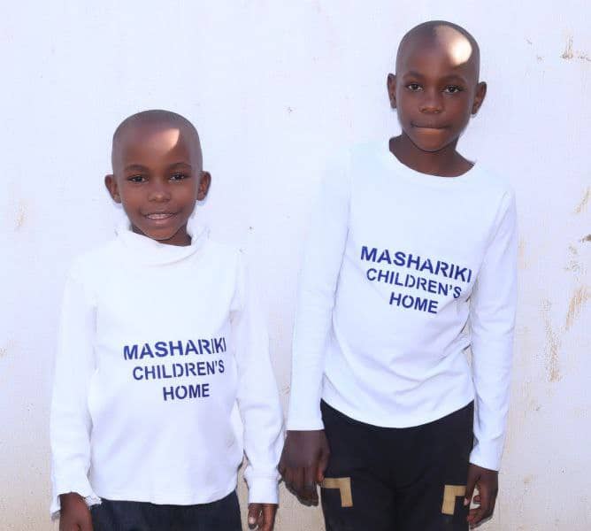 Two boys stand together wearing white and black Mashariki Children's Home branded shirts.