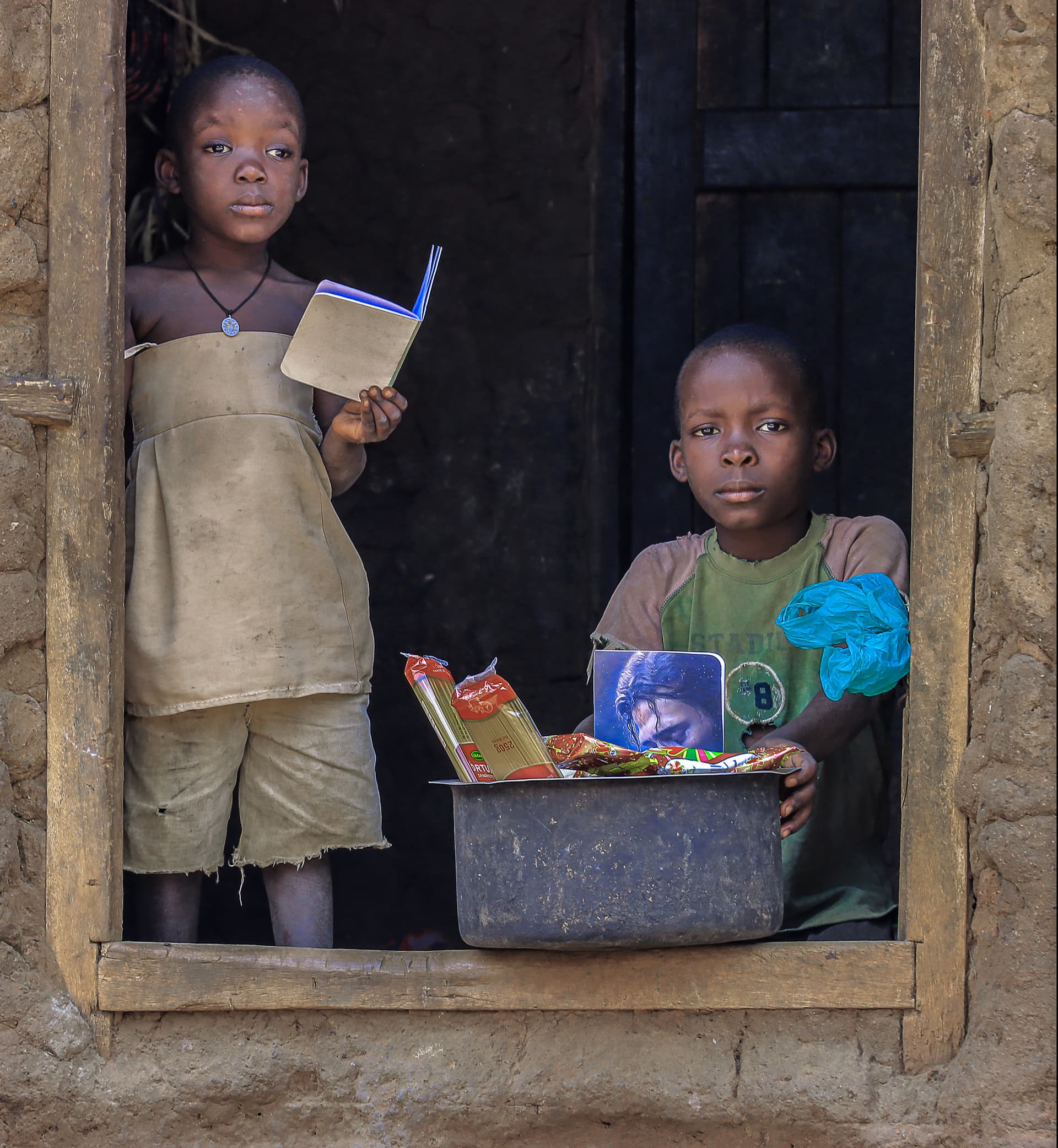 Two children stand in a mud-house doorway, one holding a food gift box beside a metal basin.