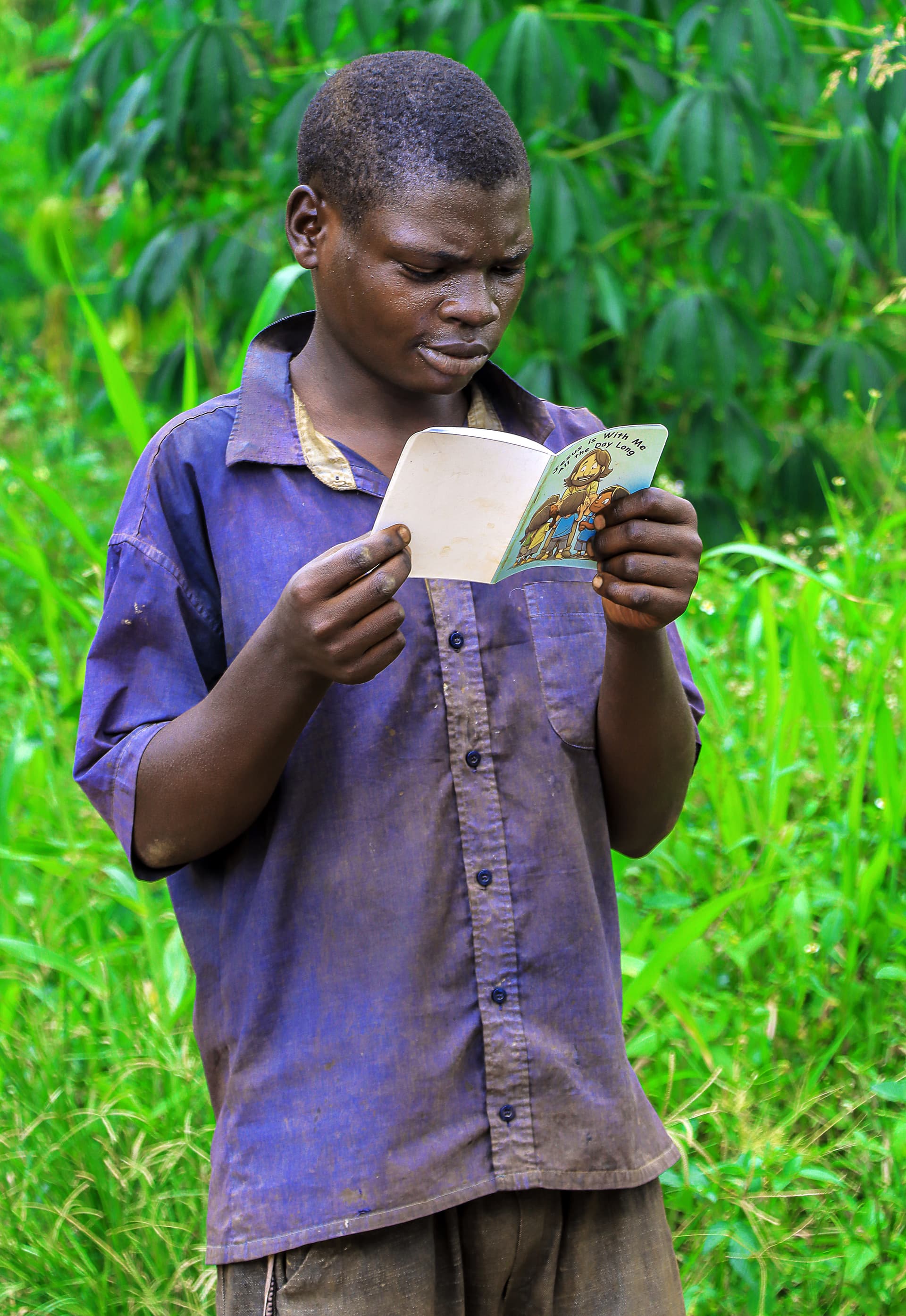 A boy in a purple striped shirt reads a booklet while standing in tall grass, identified as Israel.