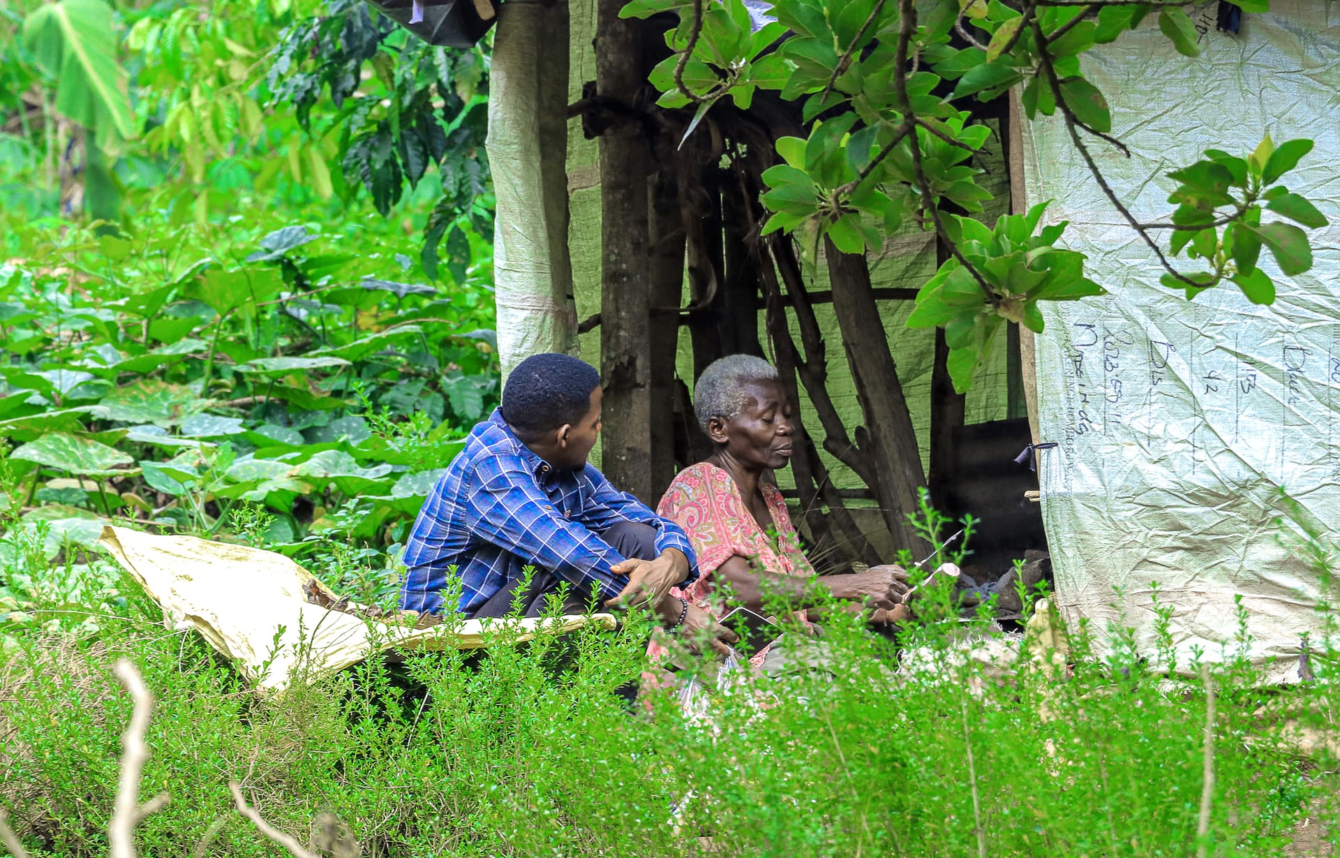 A man and an elderly woman, Gloria, sit together outdoors among greenery near a makeshift shelter.