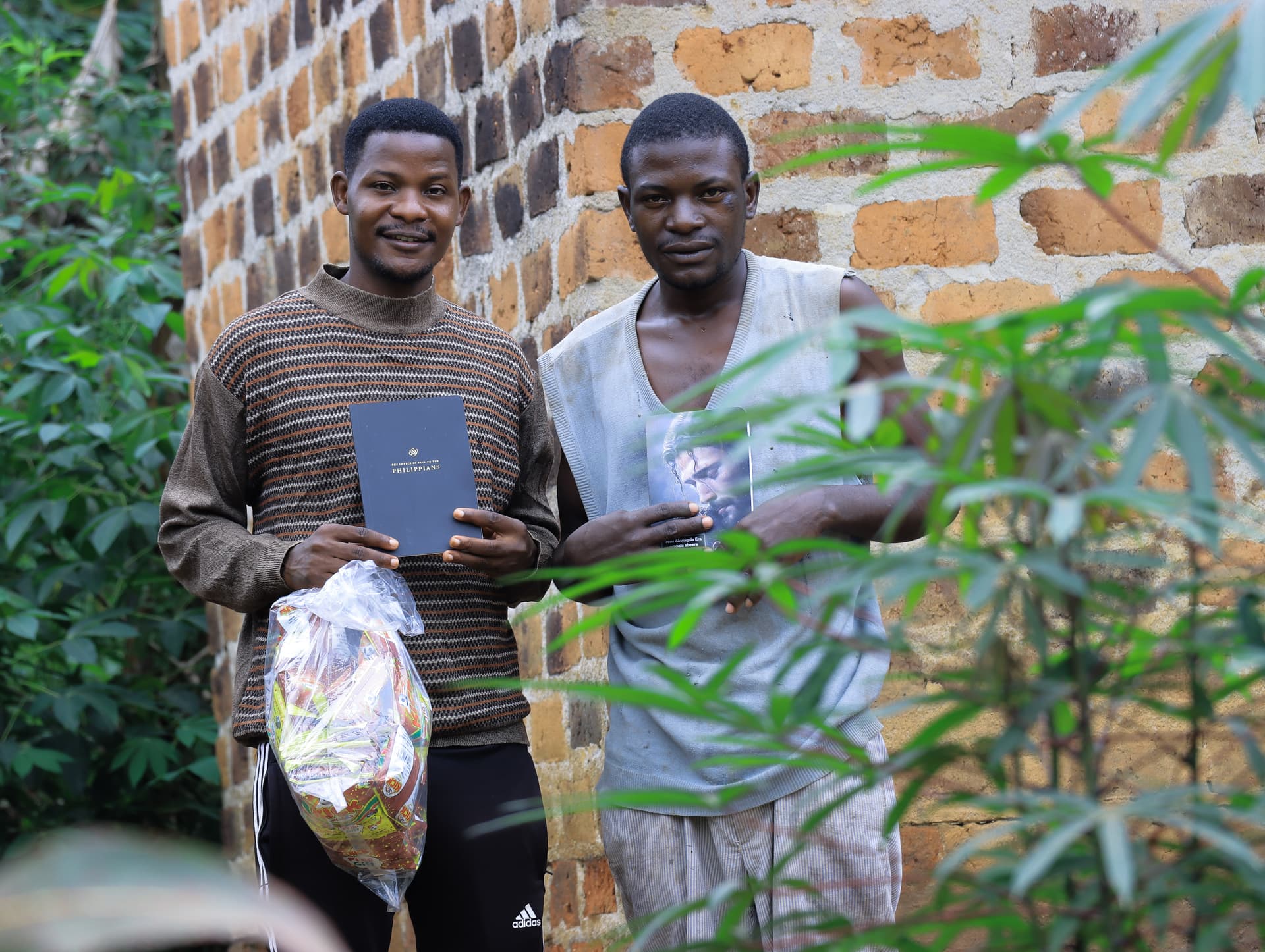 Two men stand by a brick wall, one holding a gospel tract and a plastic bag of items.