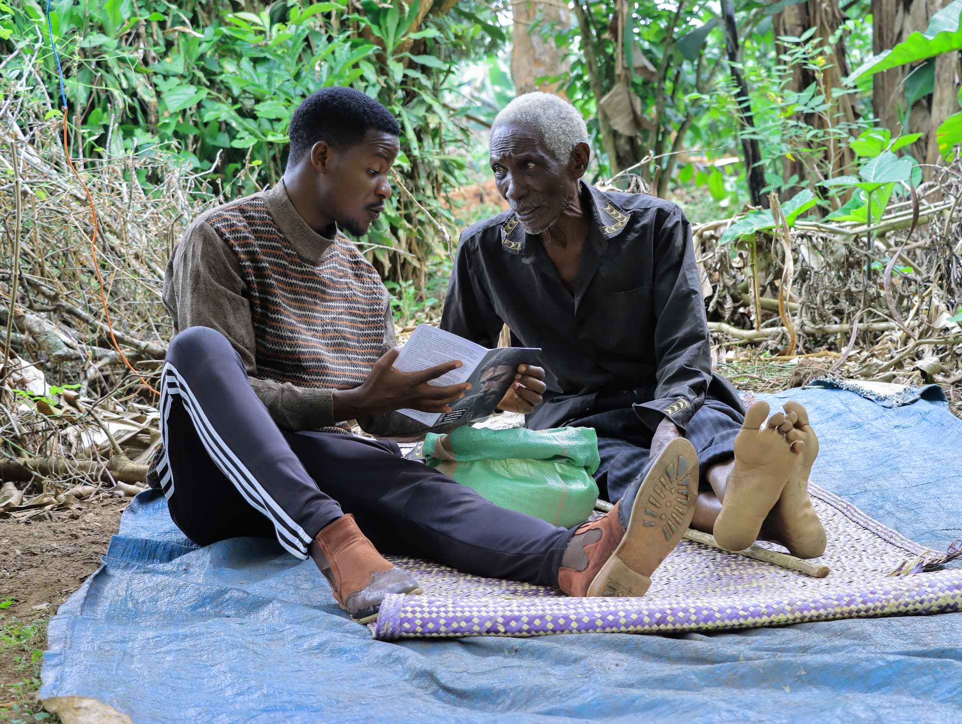 Katende Isa, an elderly man, sits on a mat outdoors talking with a younger man.