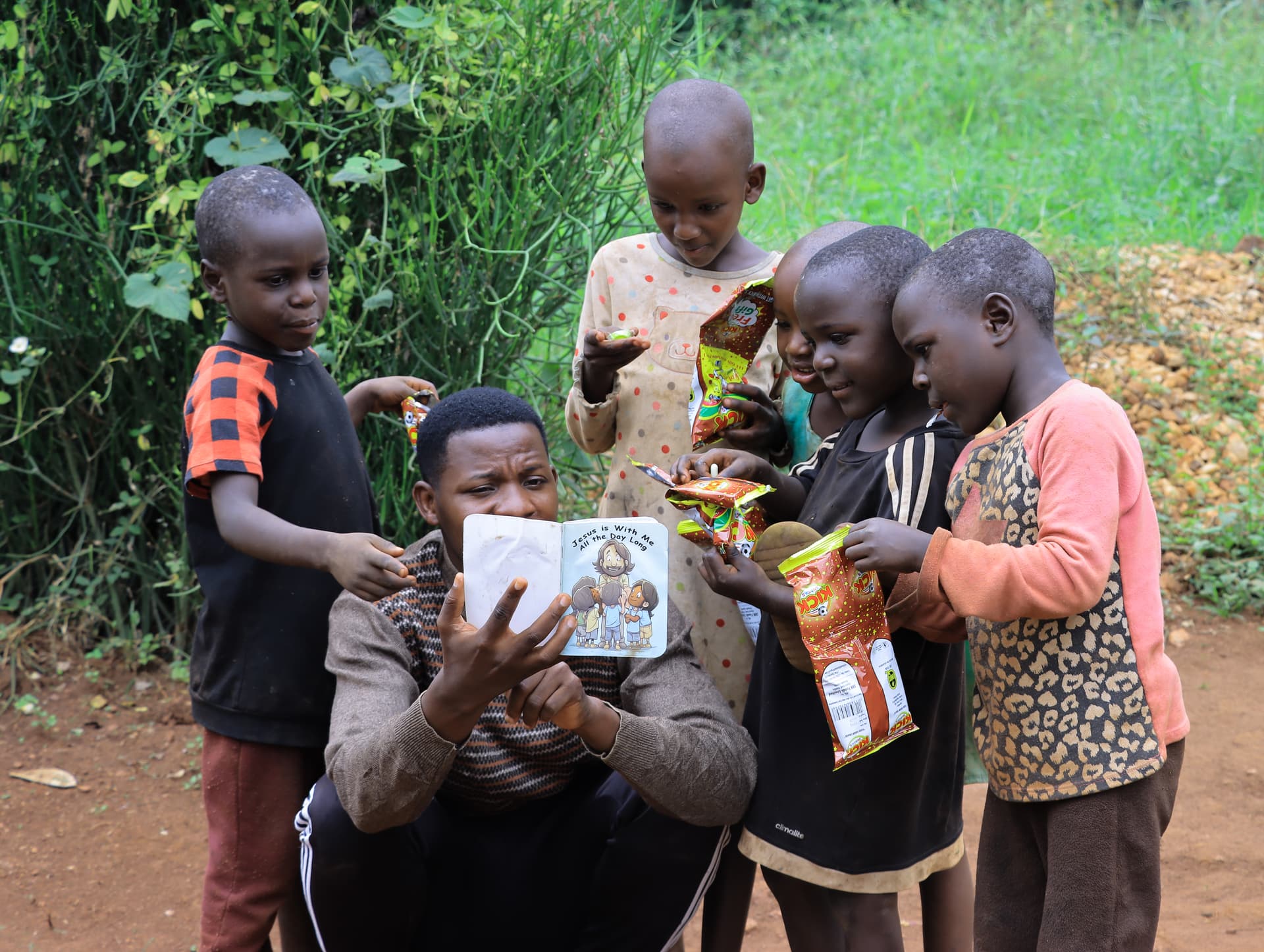 A man kneels among a group of children holding gospel tracts and food packages outdoors.