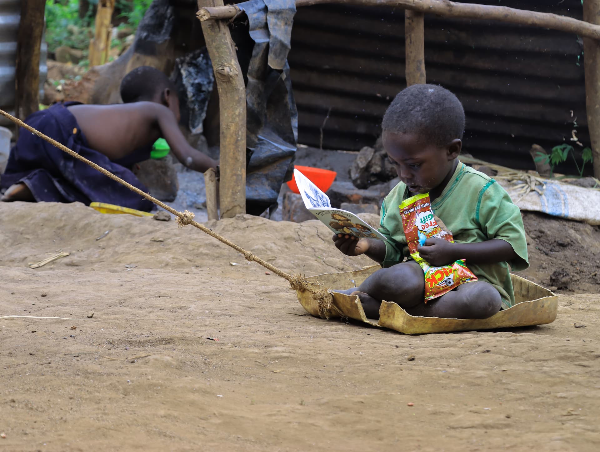 Boy sits on the ground with a homemade fishing rod near a thatched hut, a girl seated beside him.