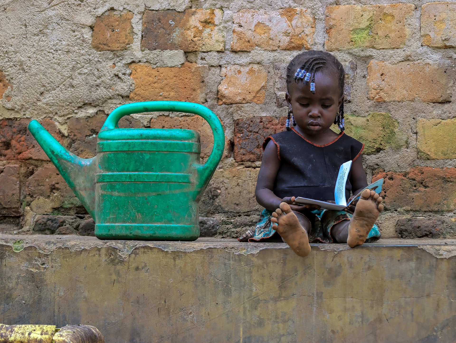 Green watering can beside a toddler seated on a low brick wall outdoors.