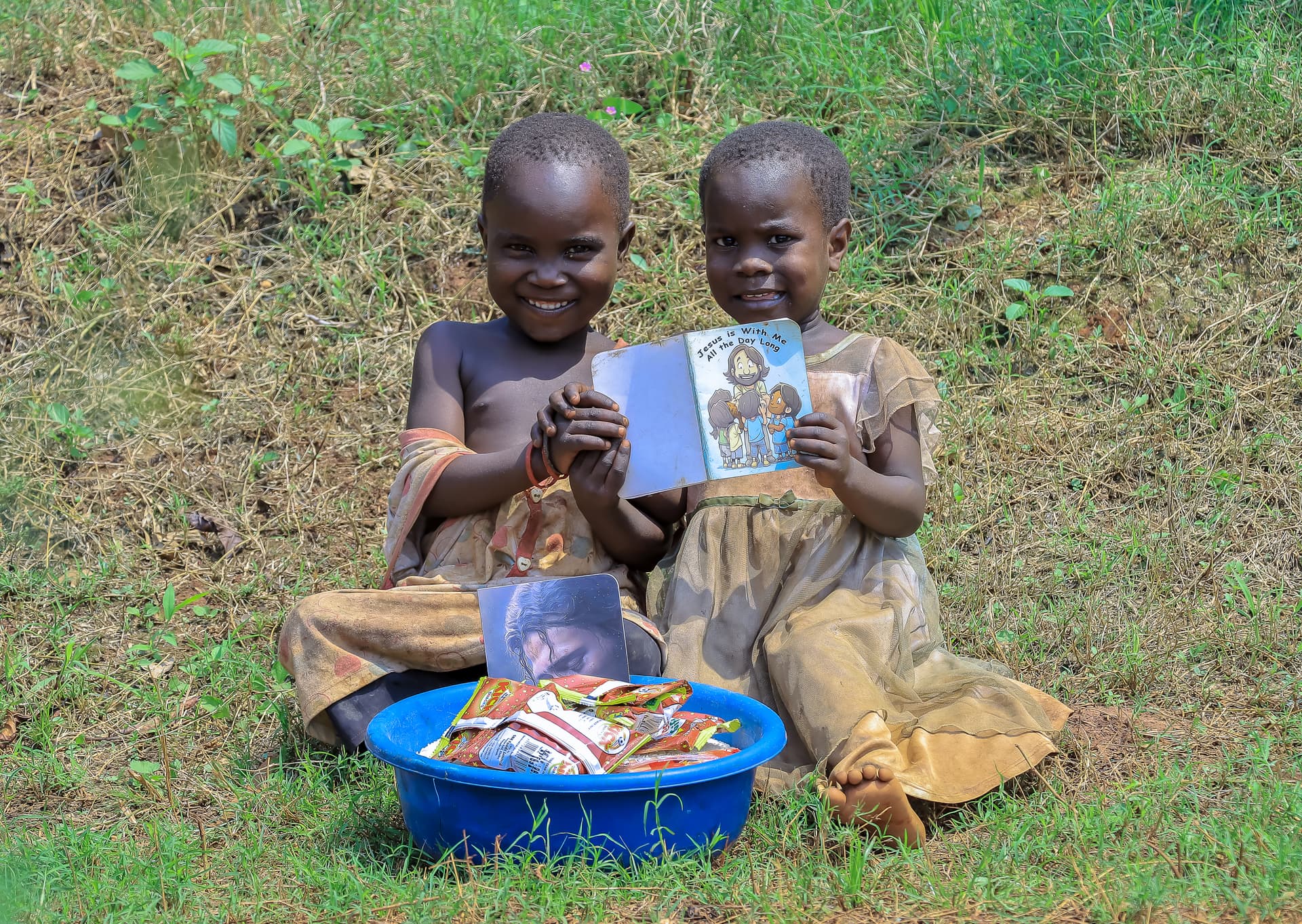 Two boys sit on the ground reading a gospel tract beside a blue basin of food gifts.